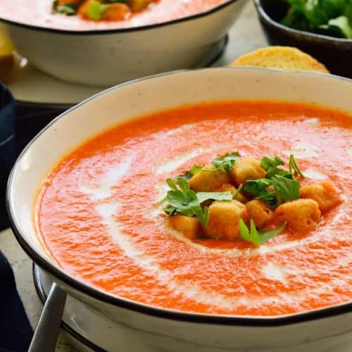 Two bowls of roasted red pepper soup garnished with croutons and chopped parsley.
