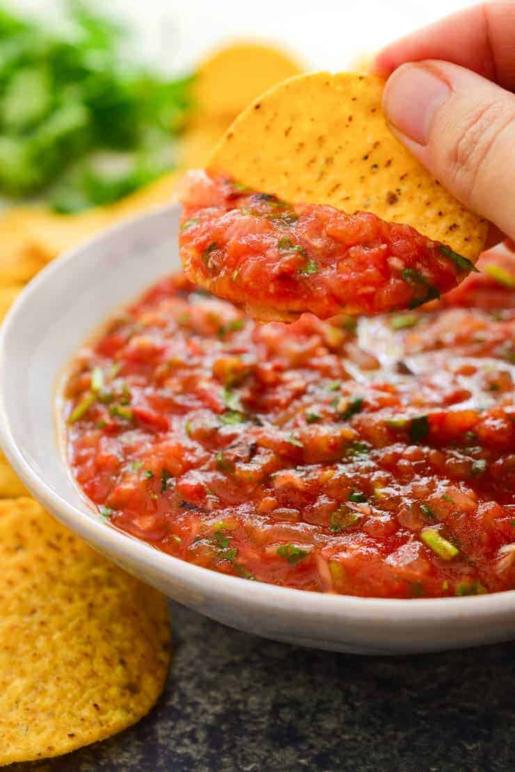 A hand holding ups a chip that has been dipped in a white bowl of roasted tomato salsa.