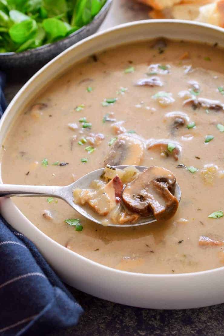 A spoon with a mushroom in a white bowl of vegan cream of mushroom soup.