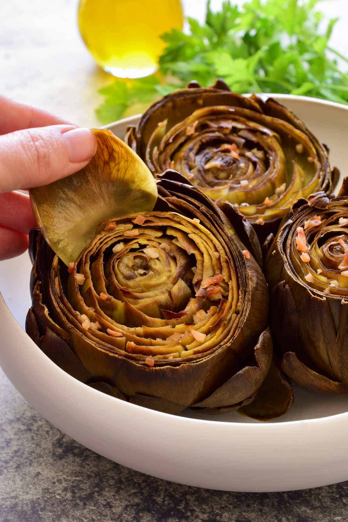 A hand pulling a leaf out of a roasted artichoke in a white bowl.