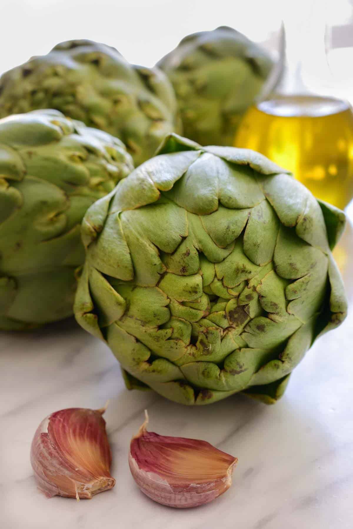 Fresh artichokes on a counter with two cloves of garlic and a jar of olive oil.