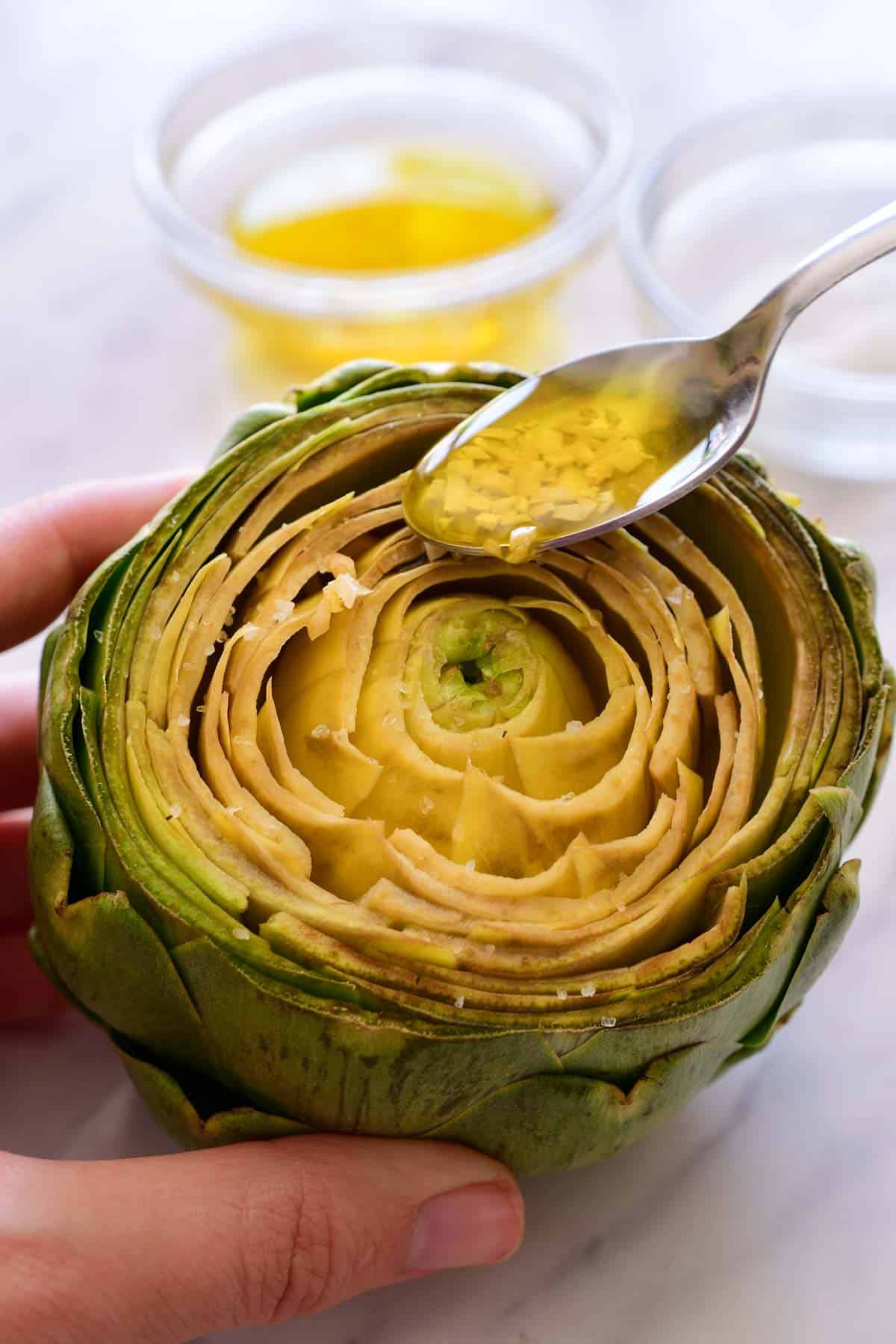 A spoon drizzling garlic oil over the top of an artichoke.