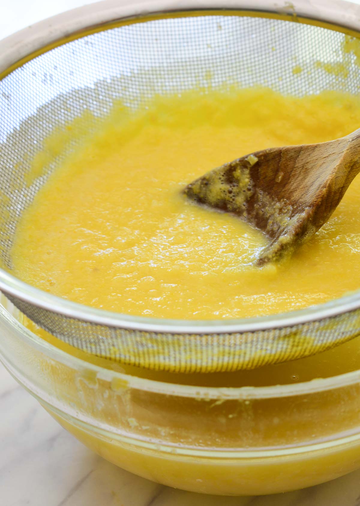 Straining the corn broth into a glass bowl through a fine mesh strainer with a wooden spoon.