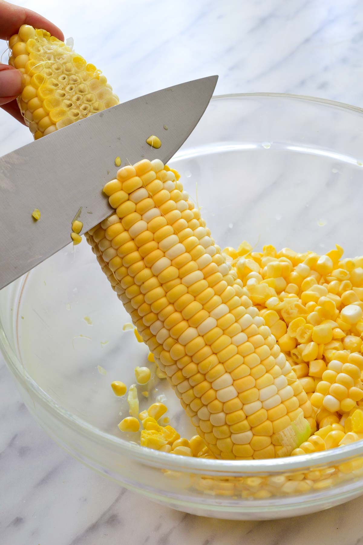 A knife cutting corn kernels off the cob over a glass bowl.