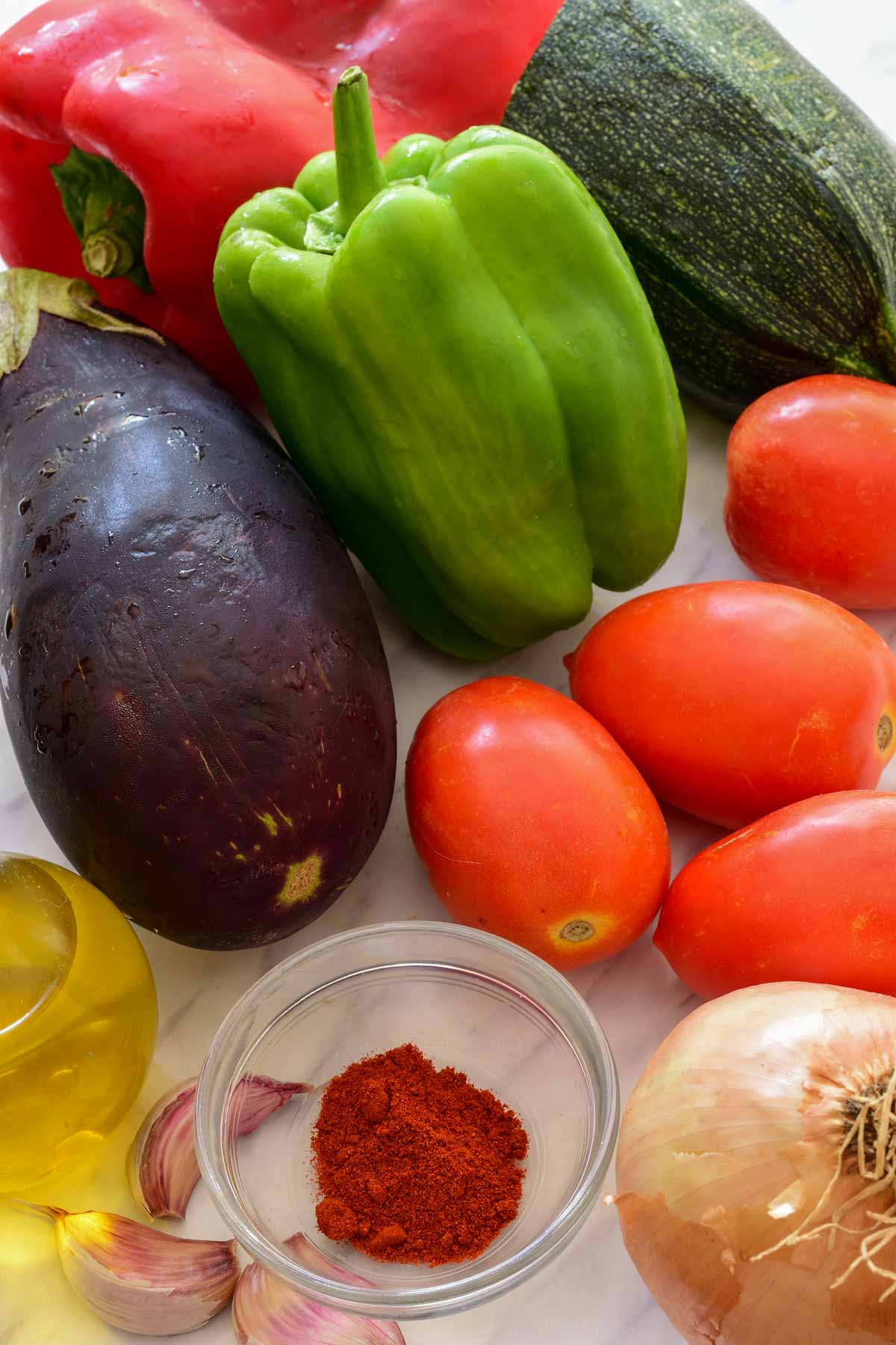 Raw ingredients on a counter: onion, tomatoes, eggplant, zucchini, green and red pepper, paprika, garlic, and olive oil.
