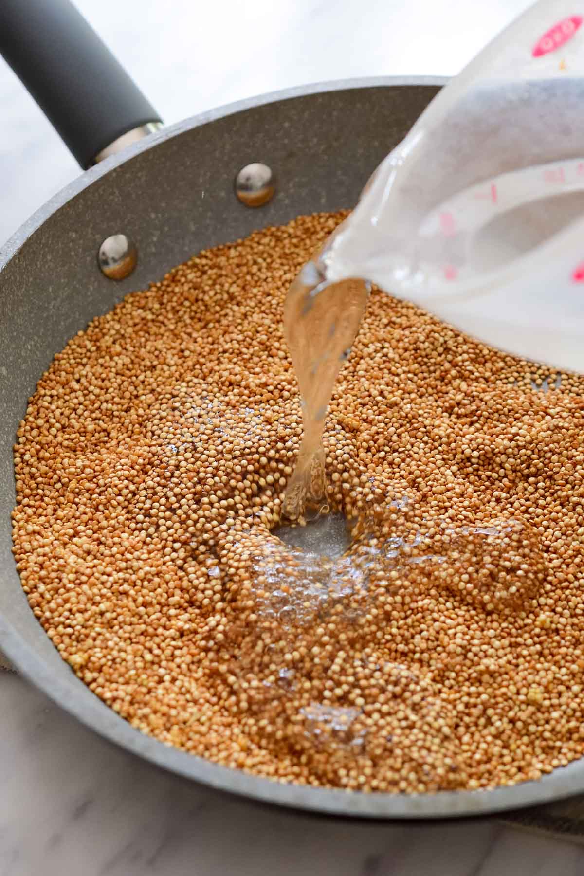 Pouring water into the grey pan of toasted quinoa.