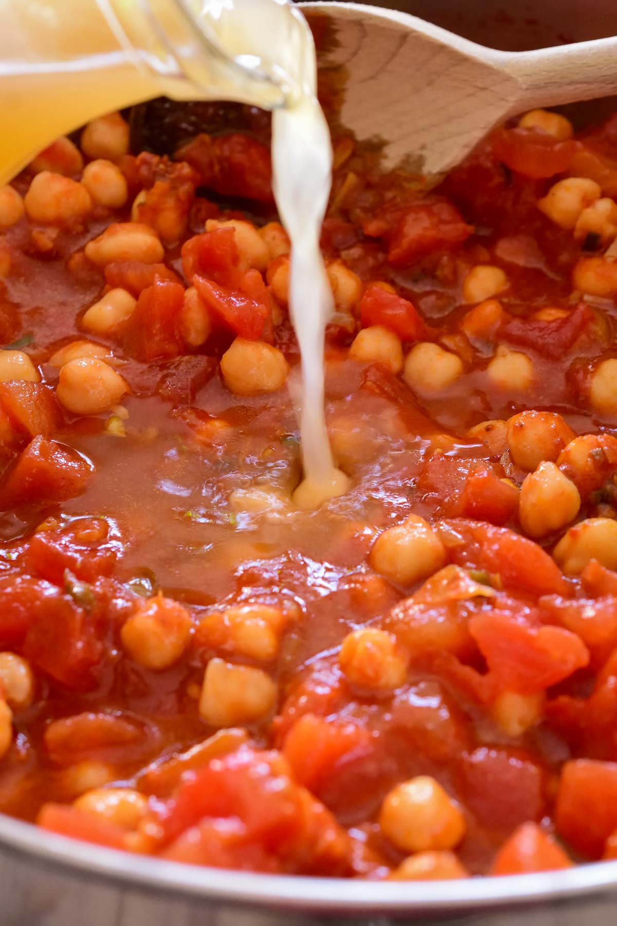 A pan of chickpeas and diced tomatoes with vegetable stock being poured in.