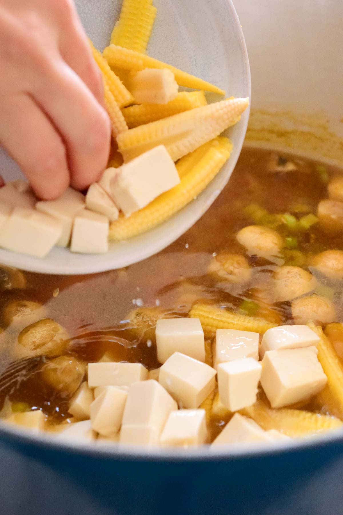 A hand pushing the tofu and baby corn into the soup pot from a small bowl.