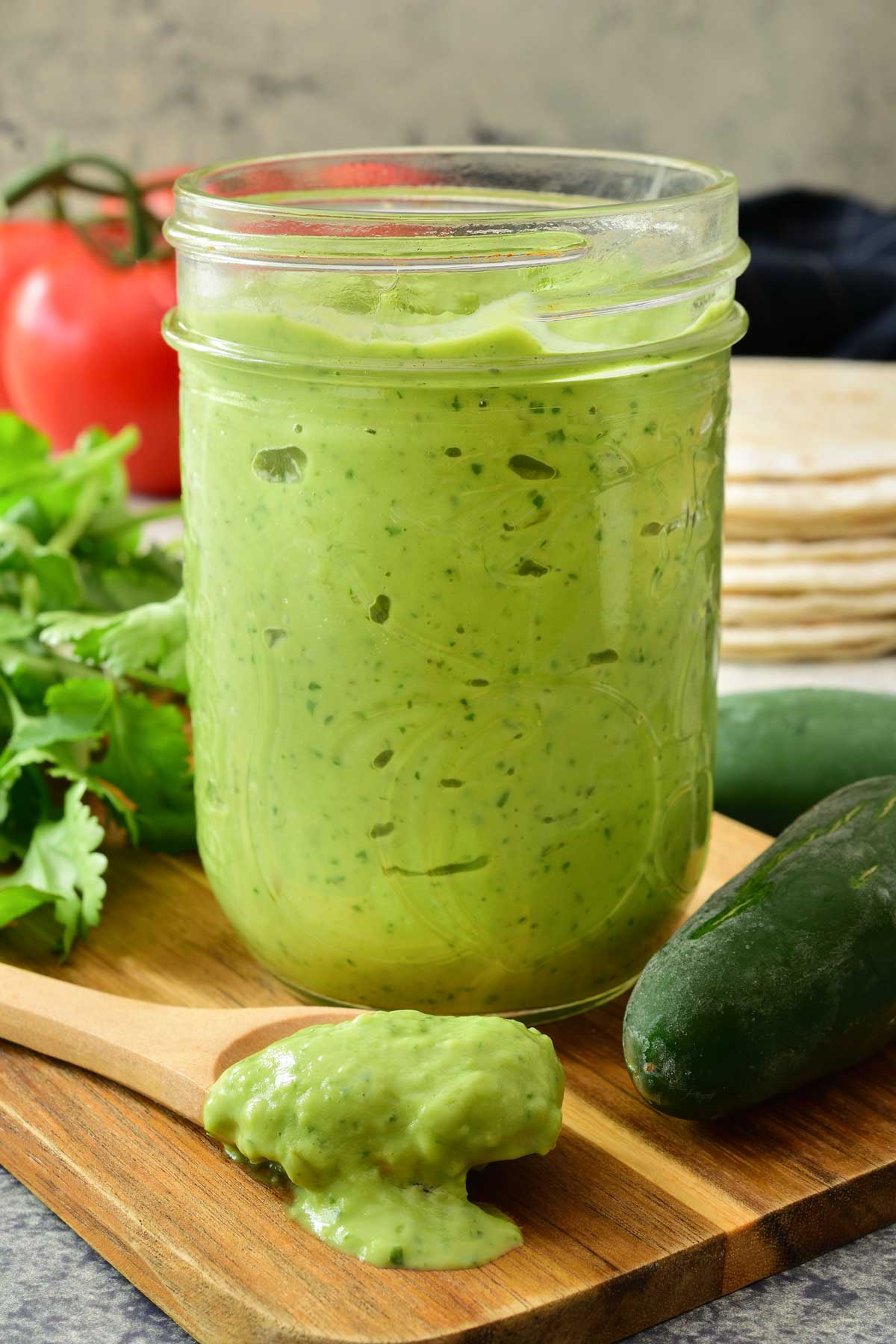 A mason jar of avocado crema on a cutting board with jalapeños, cilantro, tortillas and tomatoes in the background.