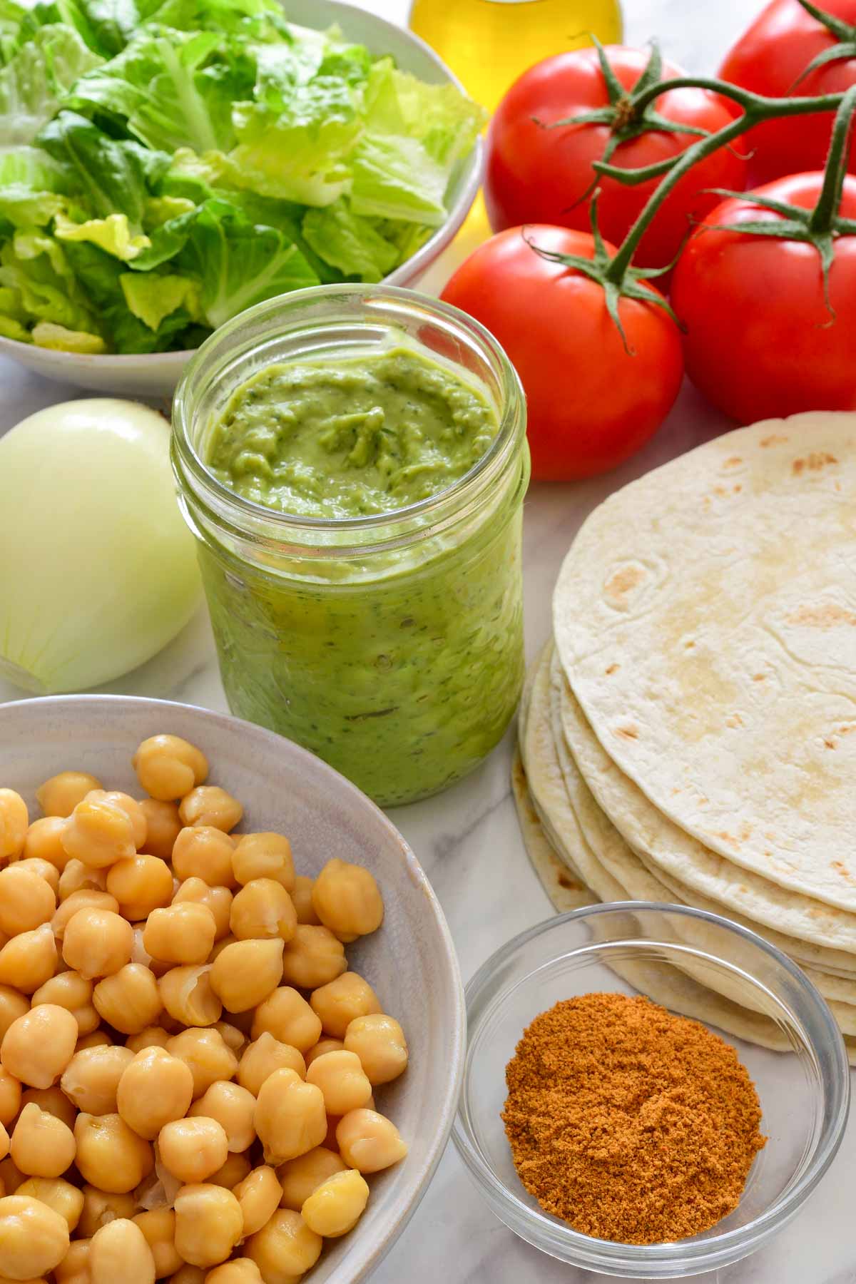 The ingredients for this recipe: a bowl of chickpeas, taco seasoning, avocado cream, tortillas, an onion, four tomatoes, a bowl of lettuce and a bottle of olive oil.