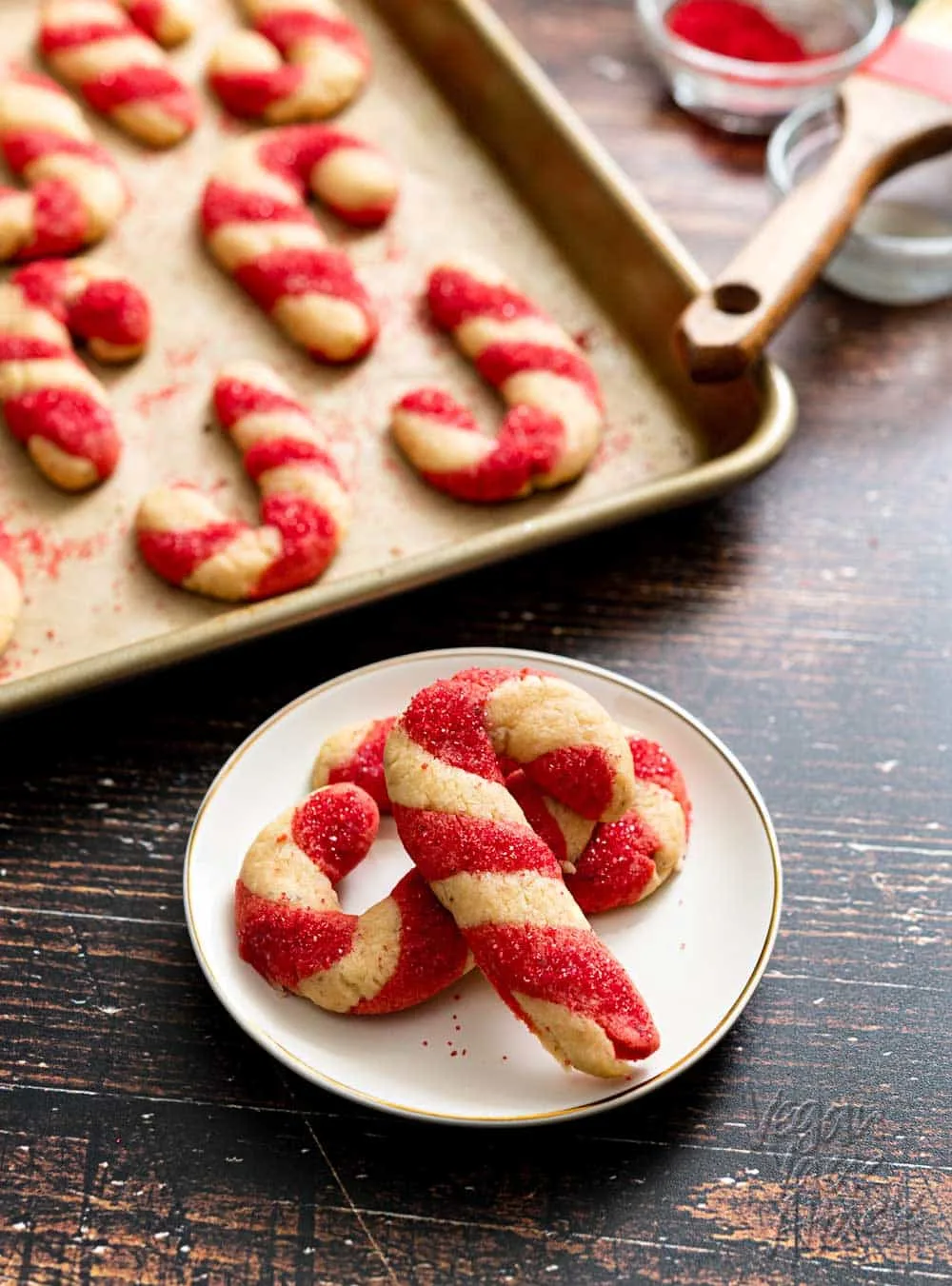 Candy cane shaped cookies on a white plate with a pan of more cookies behind.