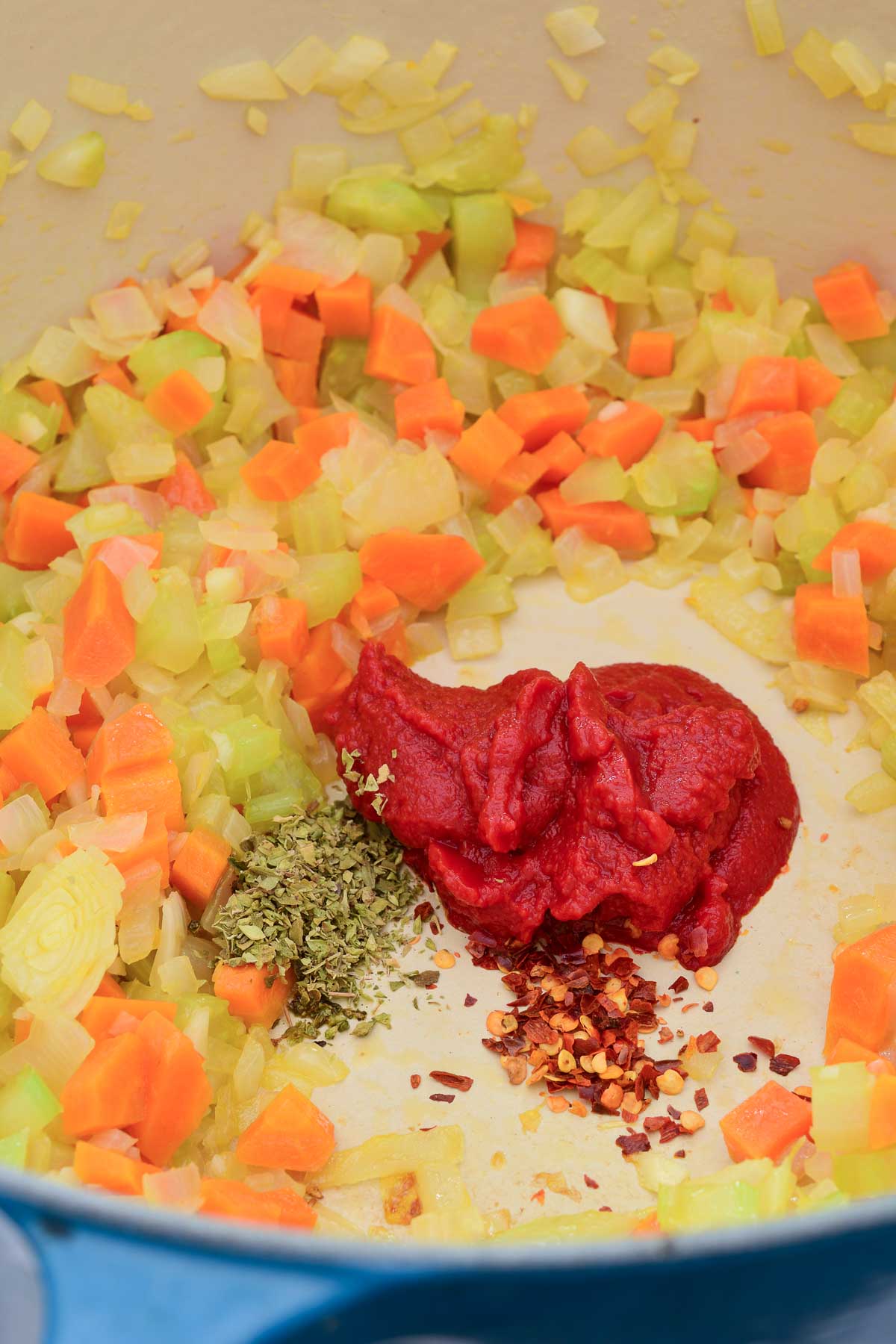 Inside a blue pot with sautéed carrots, onion and celery with a blob of tomato paste, oregano and red chili flakes in the middle.