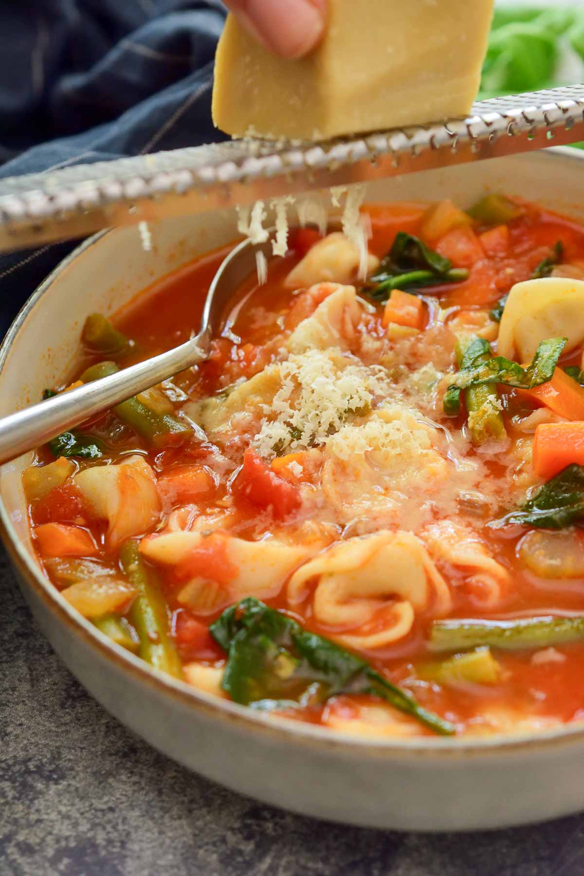 Grating vegan Parmesan cheese over a bowl of vegetarian tortellini soup.