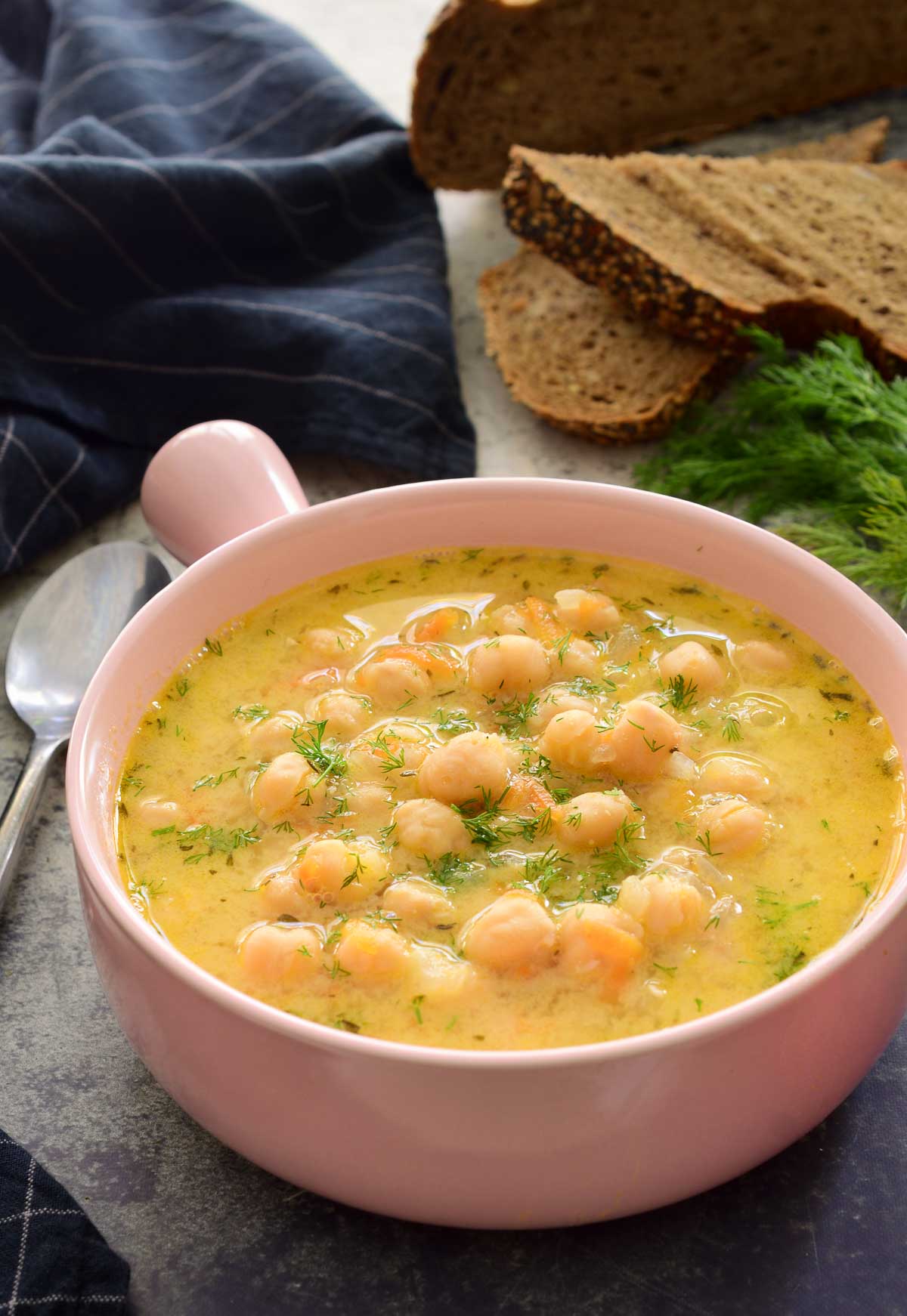 A pink bowl with a handle filled with Greek chickpea soup. A spoon, some fresh herbs, bread and a blue napkin can be seen surrounding the bowl.