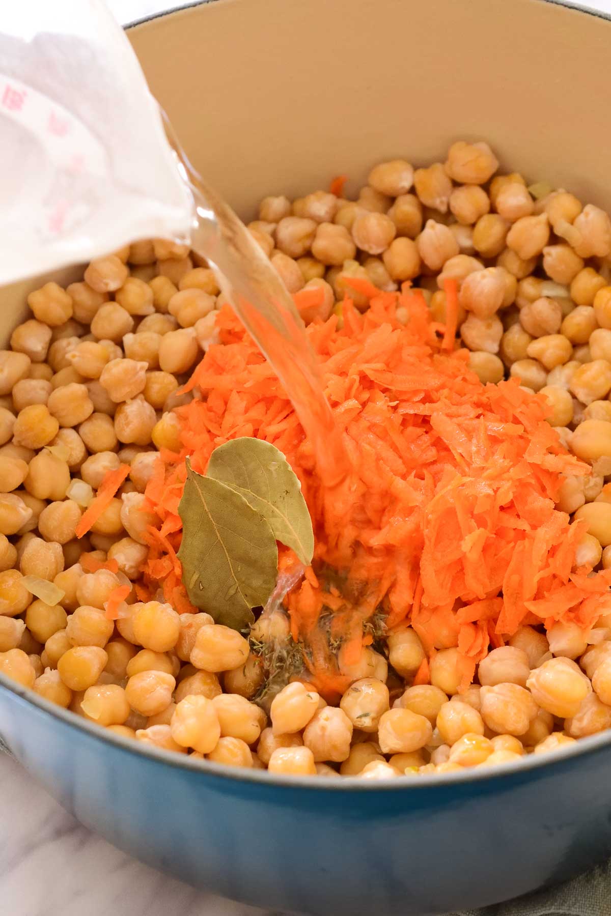 Pouring water into a blue pot filled with chickpeas, bay leaves, herbs and grated carrots.