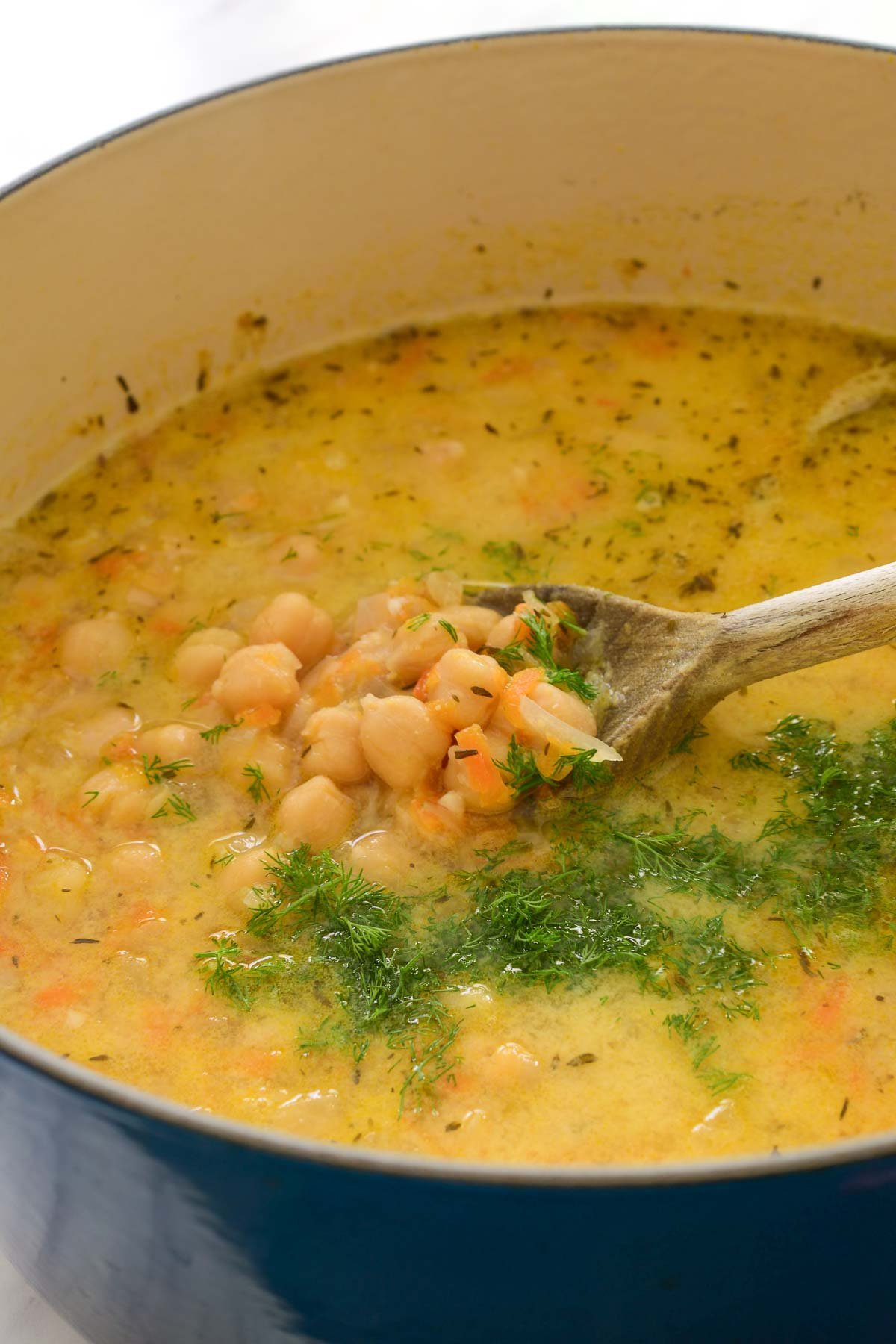 A blue pot of Greek chickpea soup with chopped fresh dill on top and a wooden spoon inside.