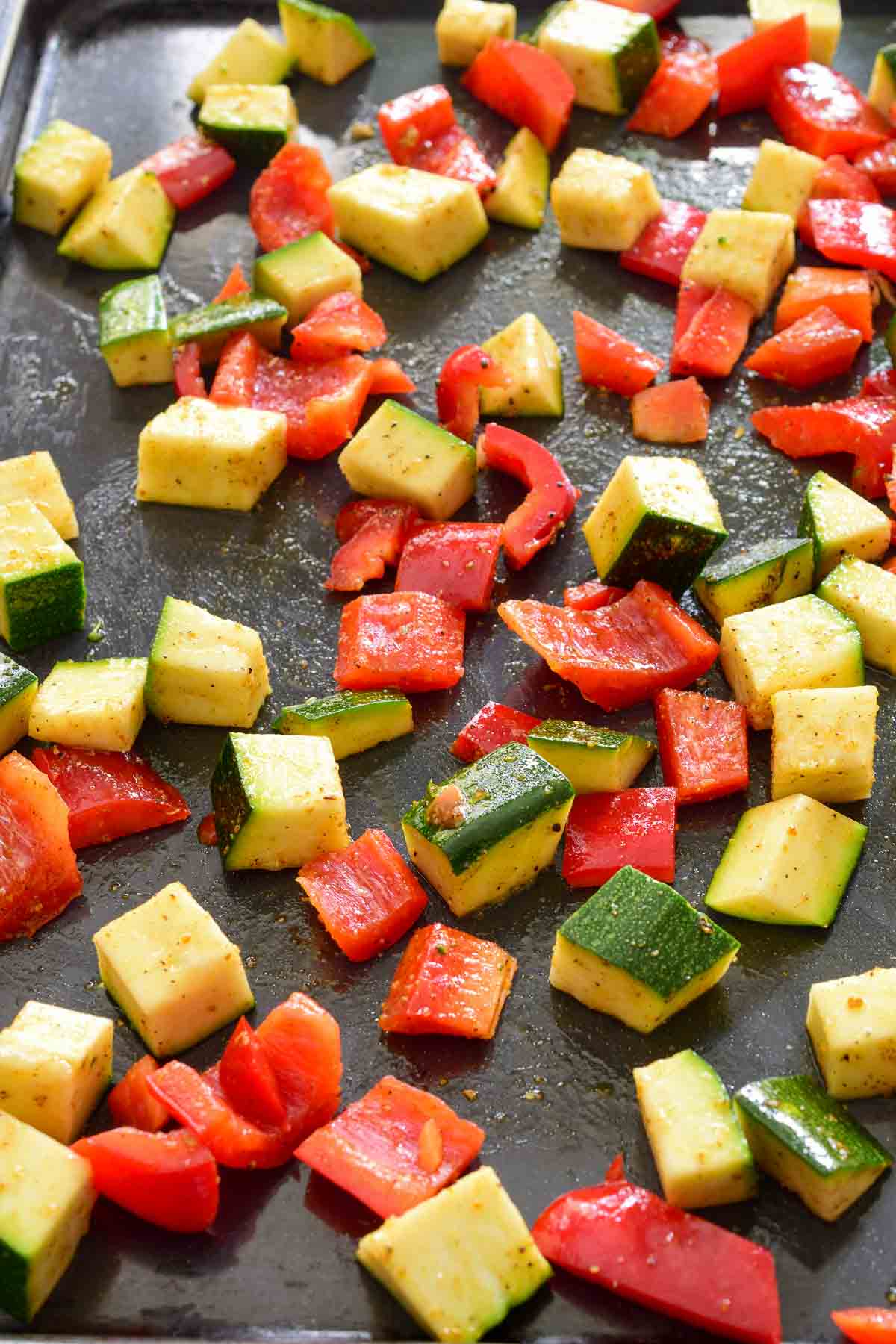 Diced zucchini and red pepper on a black baking sheet.