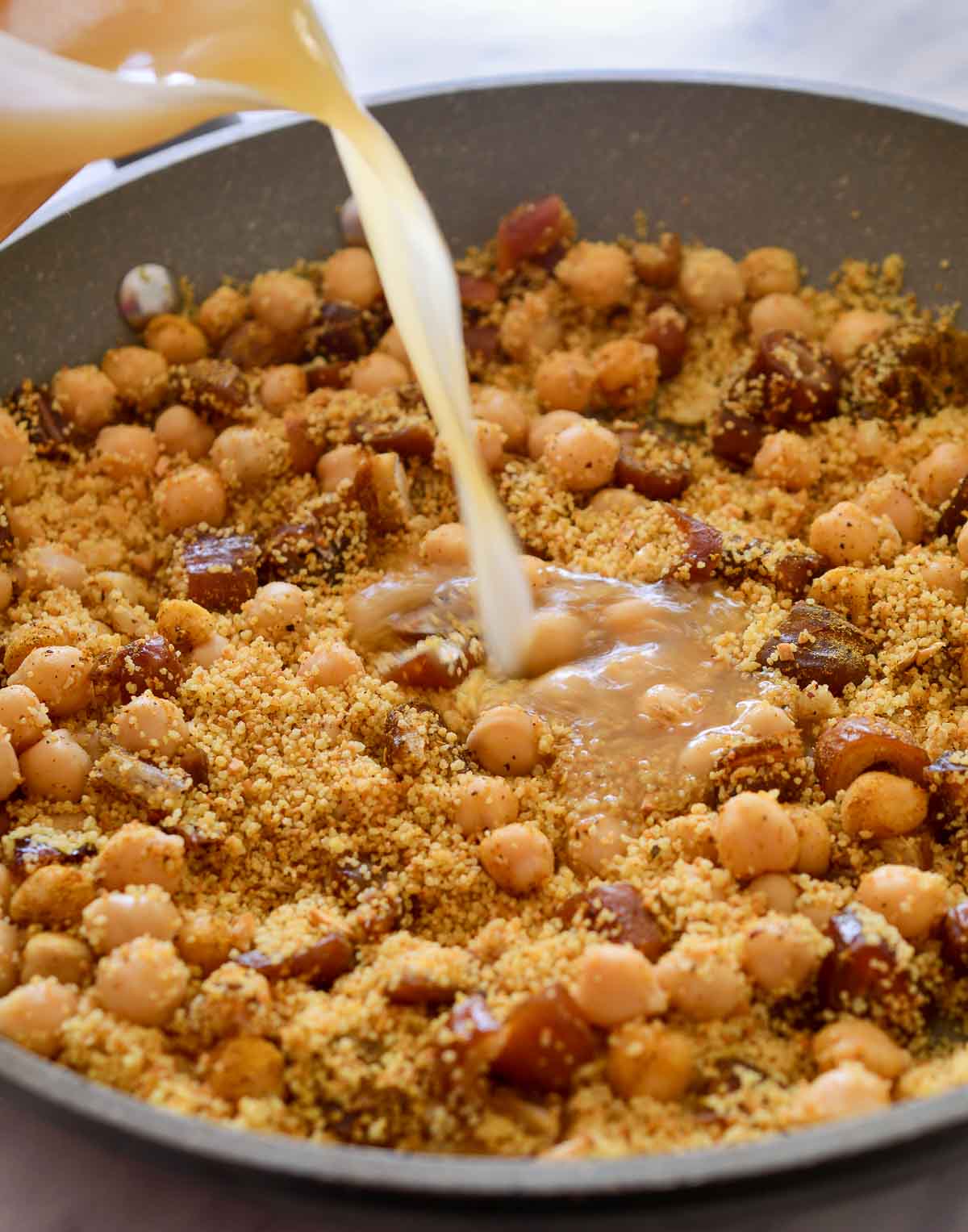 Pouring vegetable broth into the pan of mixed couscous, chickpeas and dates.