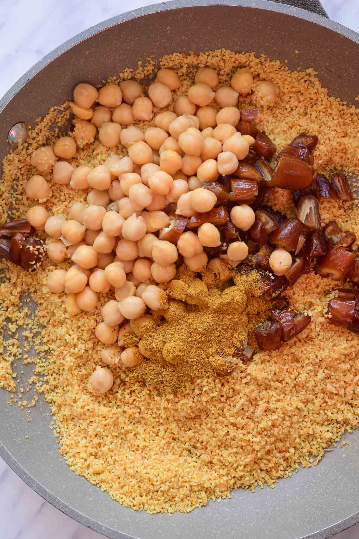 A grey pan filled with dry couscous, chickpeas, chopped dates and ras el hanout.
