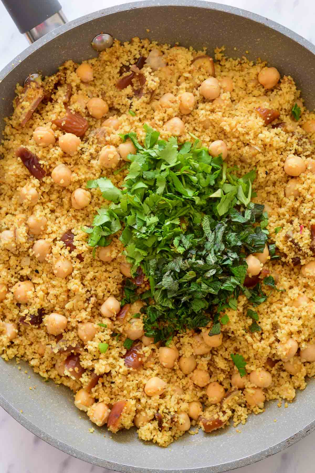 The cooked easy Moroccan couscous with chickpeas in a grey pan with a pile of fresh herbs on top.