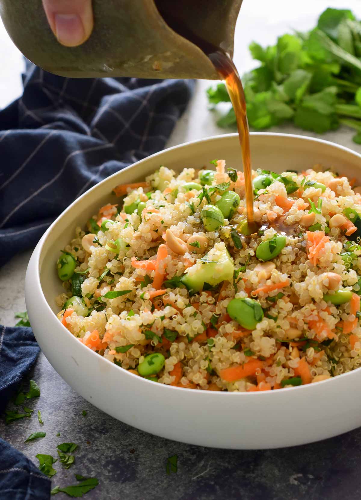 A hand holding a green jar is pouring dressing over a white bowl filled with edamame quinoa salad.