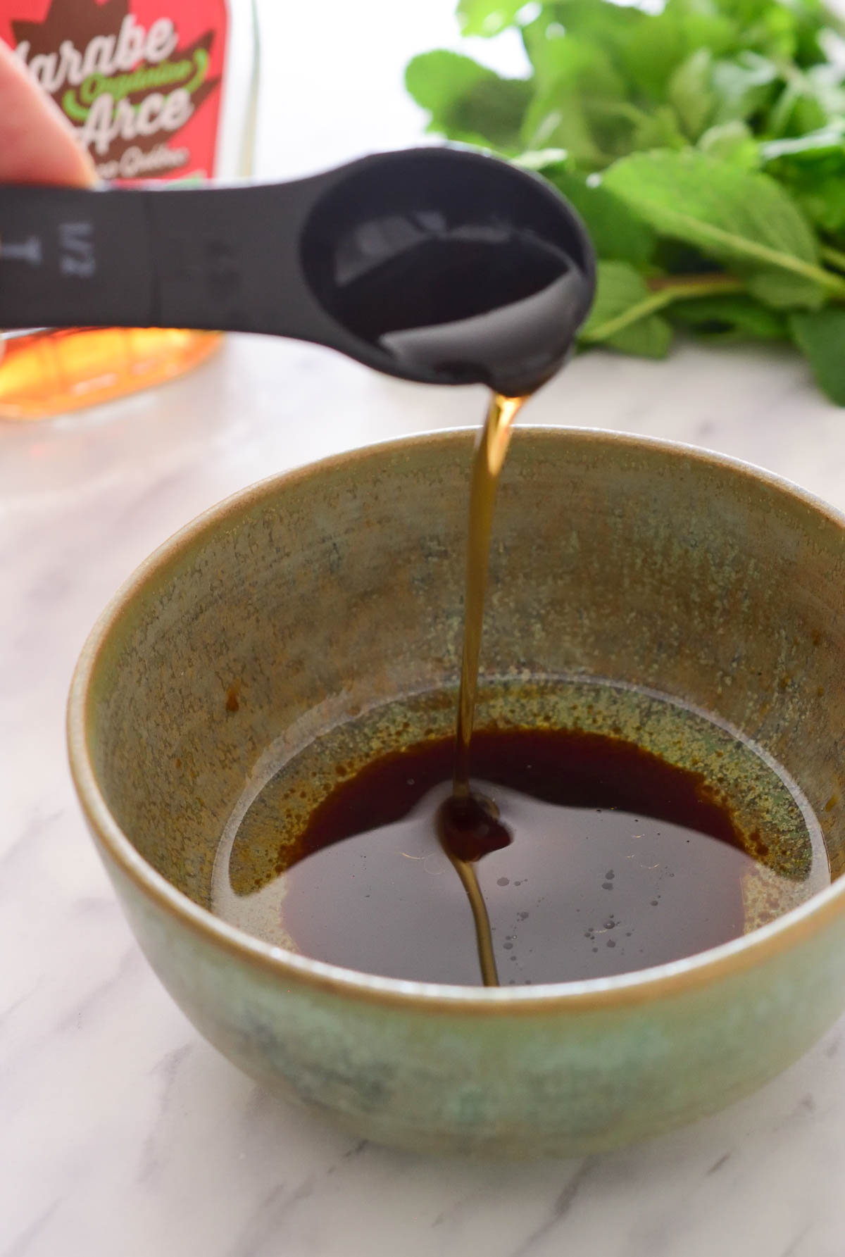 A black tablespoon pouring an amber liquid into a green bowl of salad dressing. Some herbs and a bottle are visible in the background.