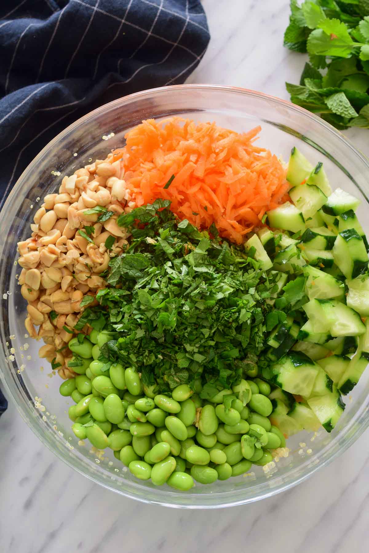 A glass bowl filled with the ingredients for edamame quinoa salad in separate piles. A blue napkin and some herbs are visible in the background.
