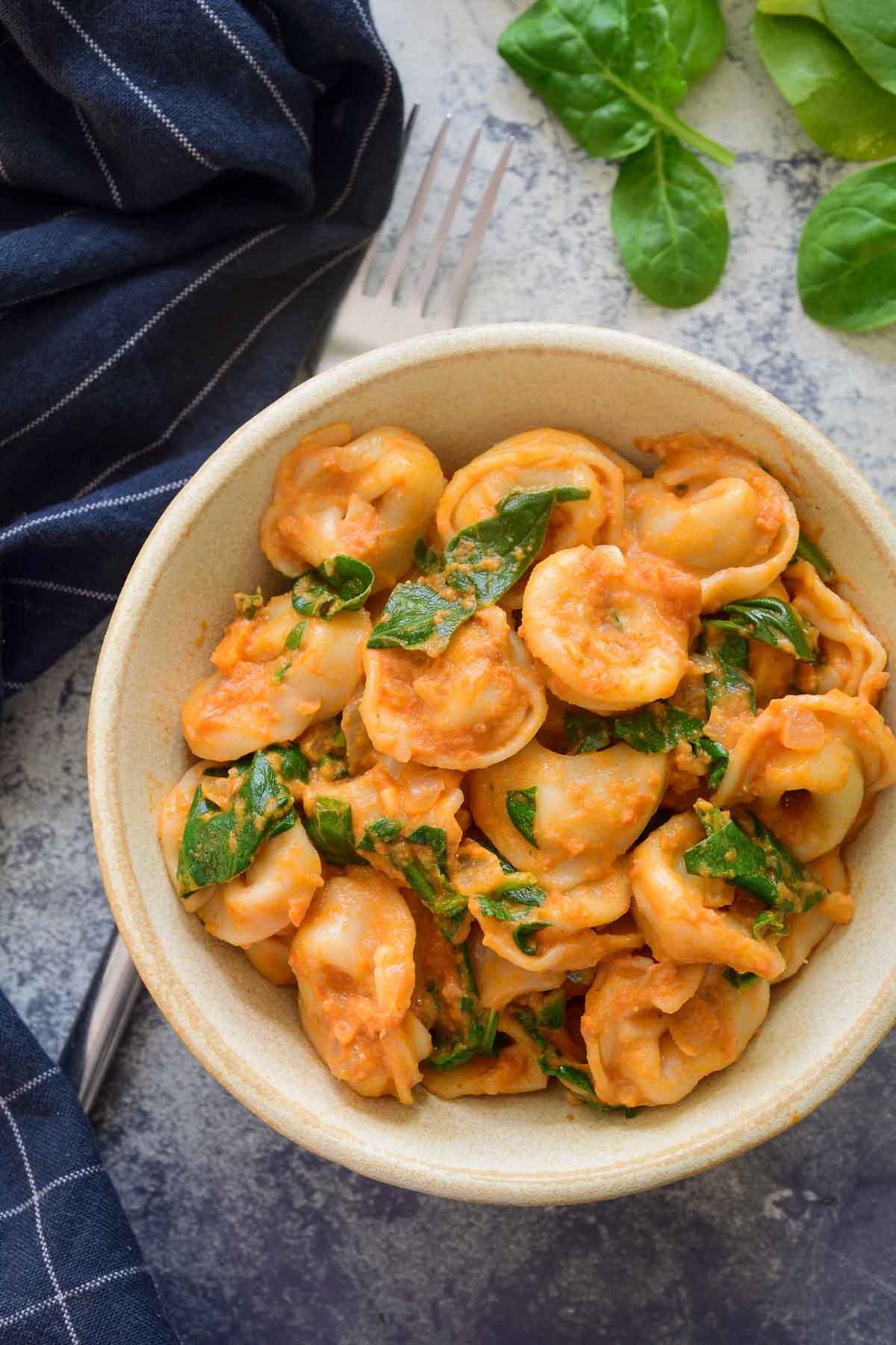 A beige ceramic bowl filled with spinach tortellini on a blue stone countertop. A blue napkin and spinach leaves surround the bowl.