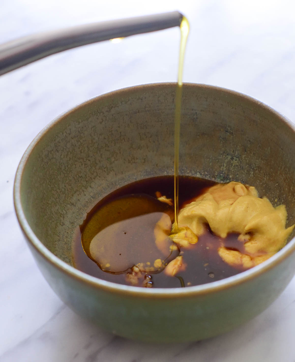 A green ceramic bowl with vinegar, maple syrup and Dijon mustard. A silver spout is pouring olive oil into it.