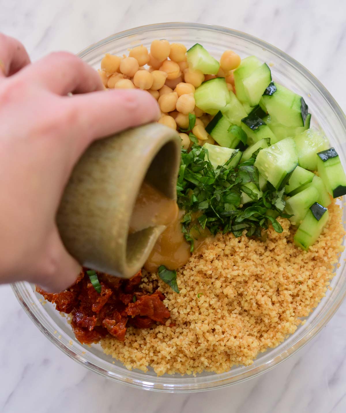 A hand holding a green ceramic jar and pouring vinaigrette over the couscous chickpea salad.