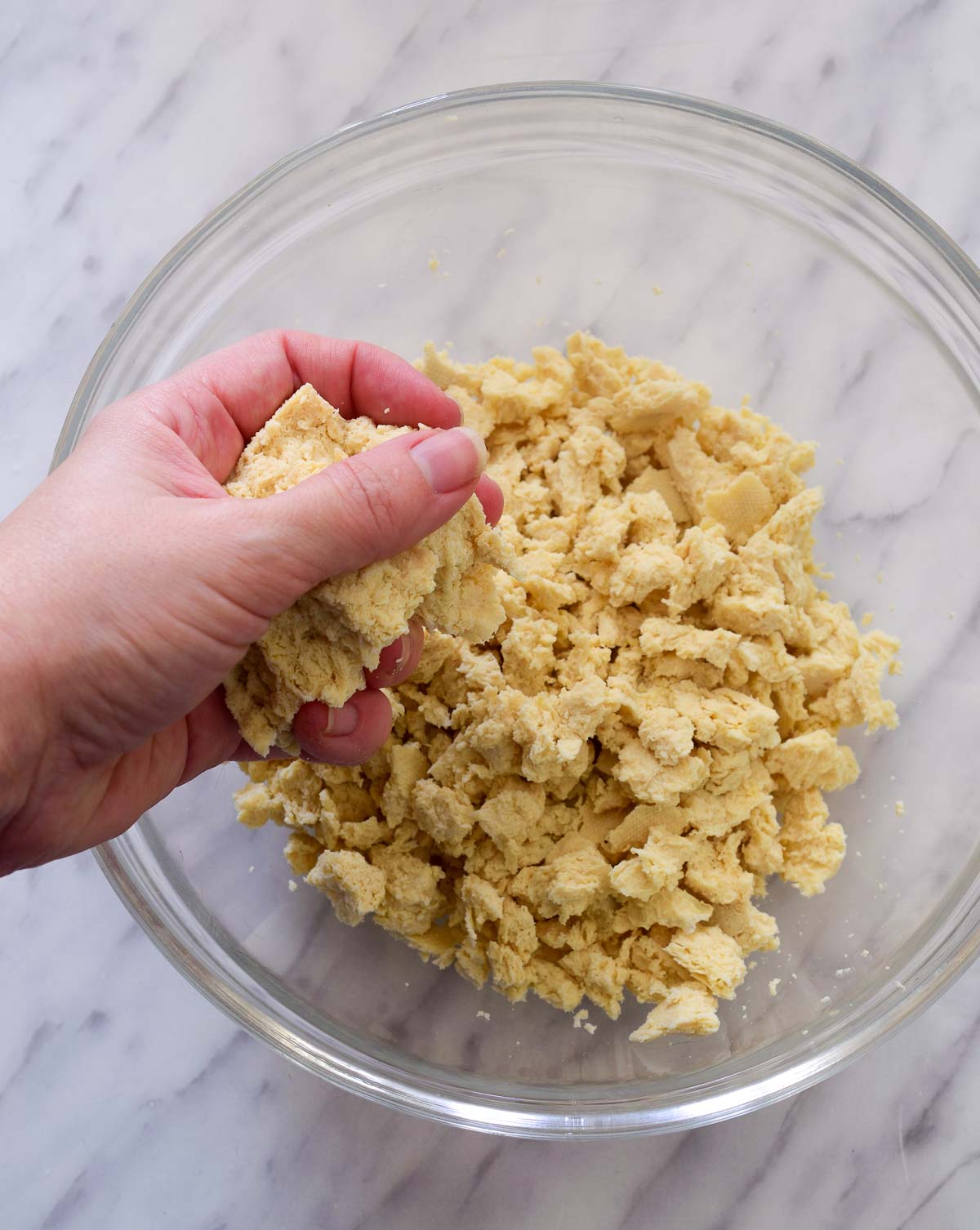 A hand crumbling a block of tofu into a glass bowl.