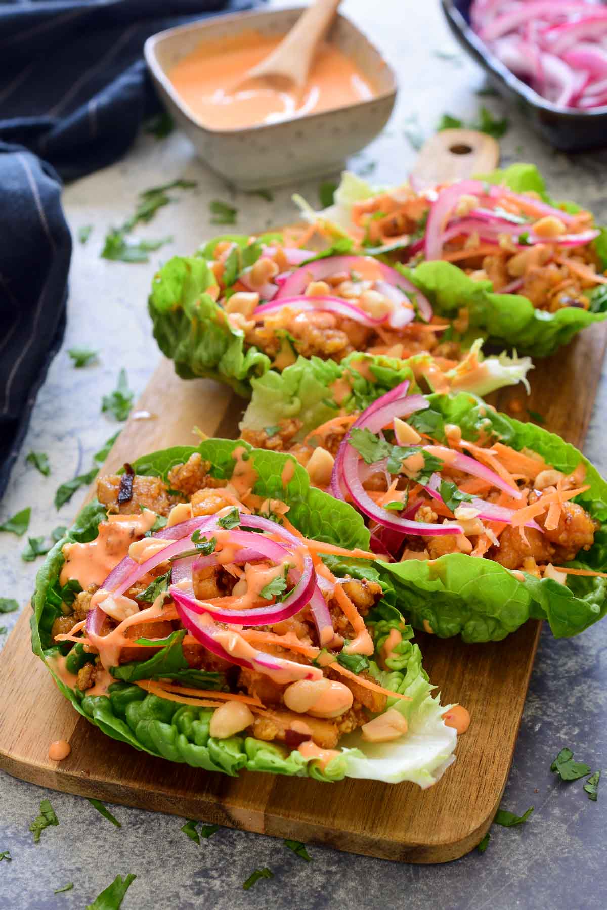 A 45-degree angle of four tofu lettuce wraps arranged on a wooden cutting board. The sauce and pickled red onions can be seen in the background.