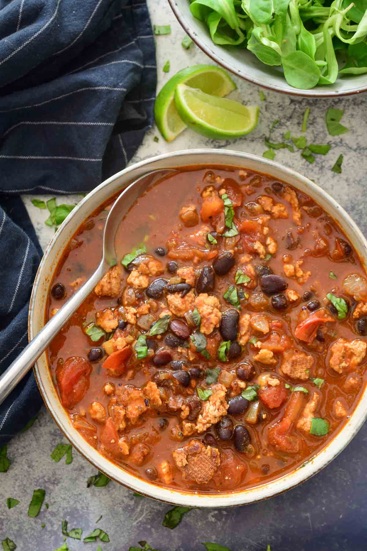 A grey ceramic bowl filled with tofu chili with a metal spoon in it. In the background are a blue striped napkin, lime slices and a second bowl filled with leafy greens.