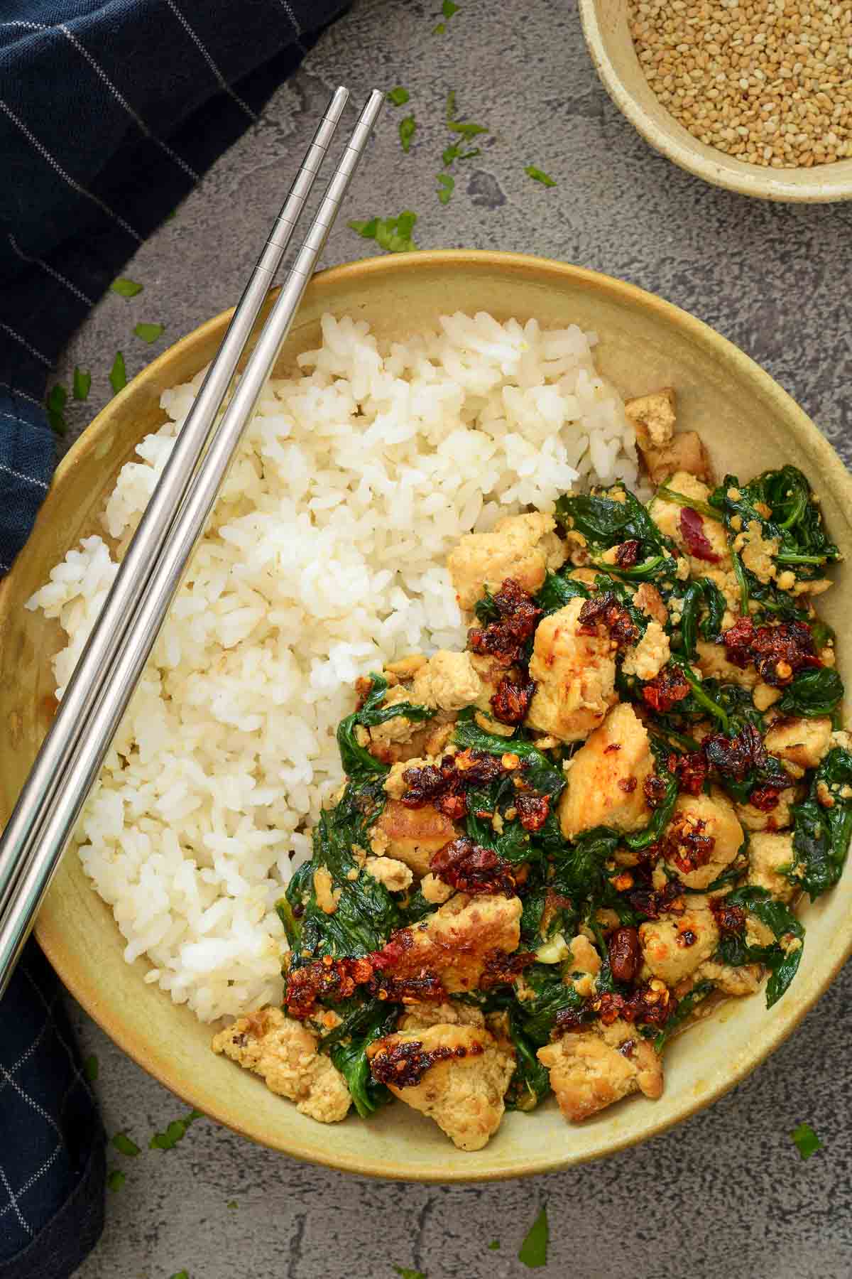 A ceramic bowl half filled with rice, half filled with spinach and tofu stir fry. A pair of silver chopsticks sits on the side of the bow.