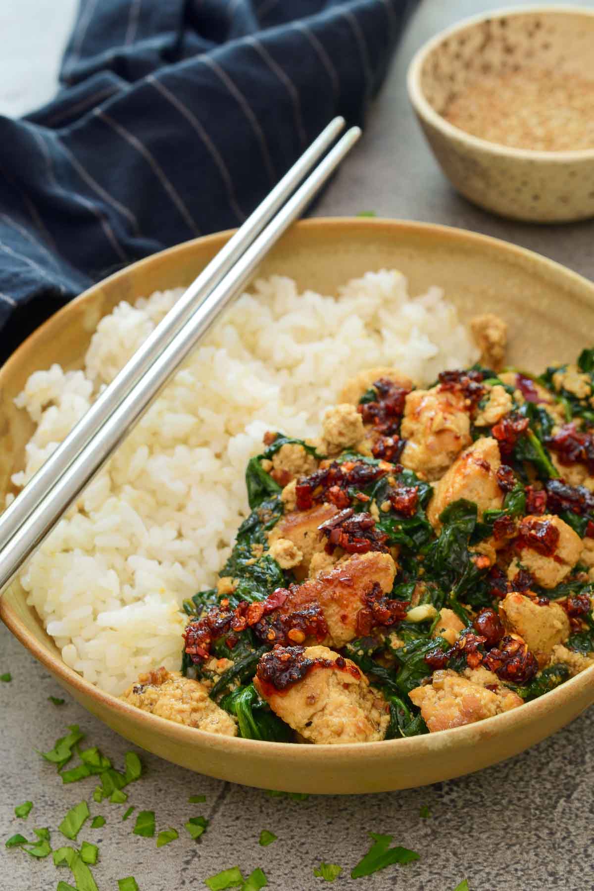 A beige ceramic bowl half filled with rice and half filled with spinach and tofu stir fry. A pair of silver chopsticks sits on the side of the bowl.