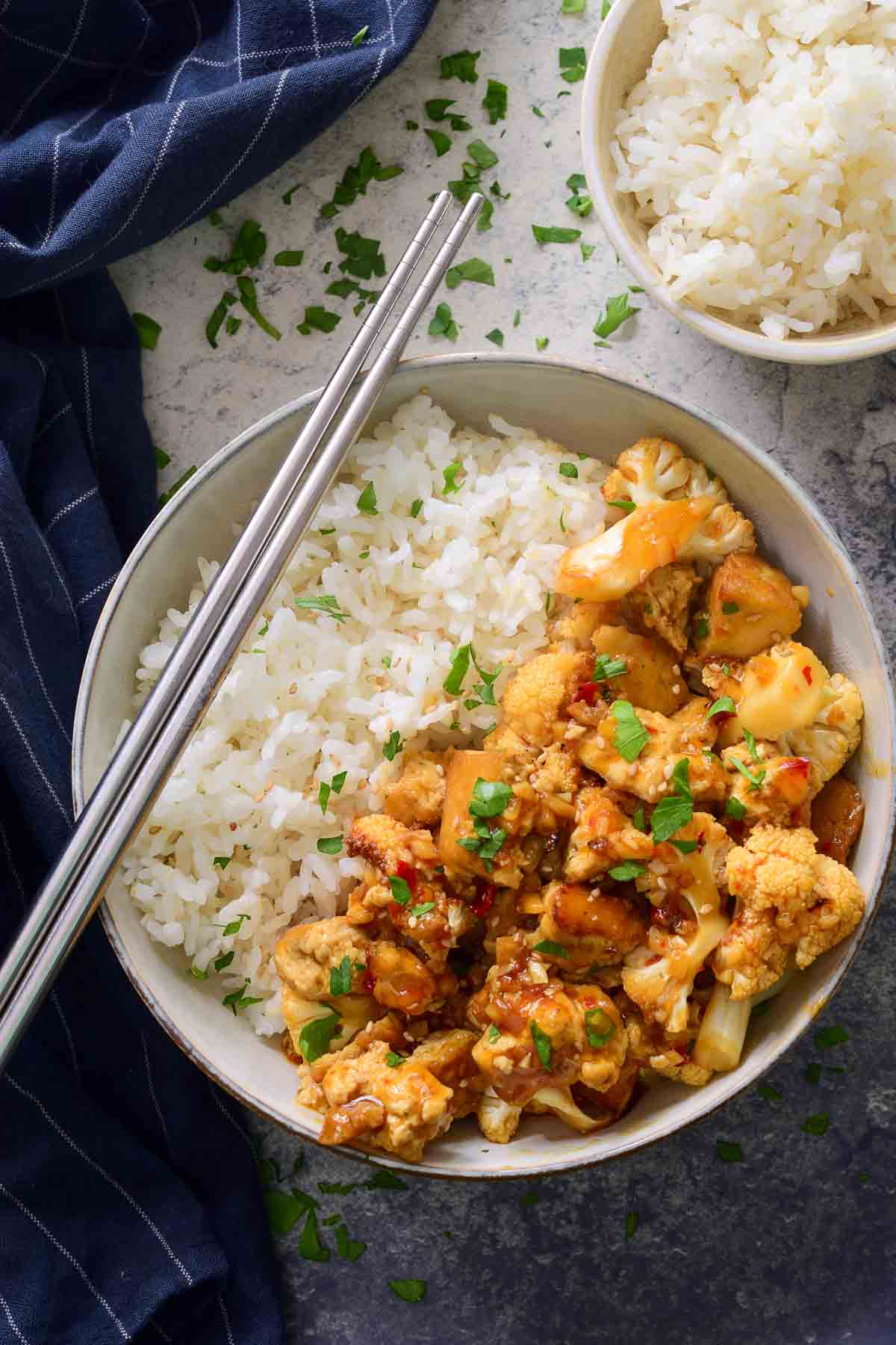 A ceramic bowl half filled with rice and half filled with tofu stir fry. A pair of silver chopsticks rests on the side of the bowl.