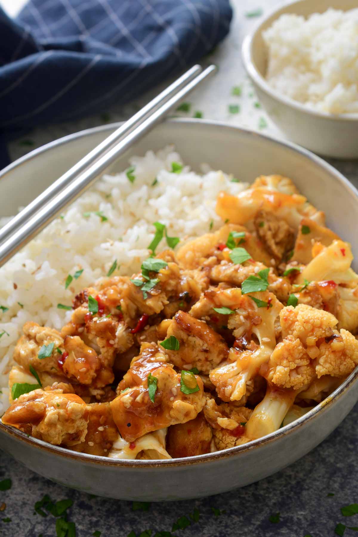 A close-up image of the tofu stir fry in a grey bowl with rice and metal chopsticks balanced on the side of the bowl.