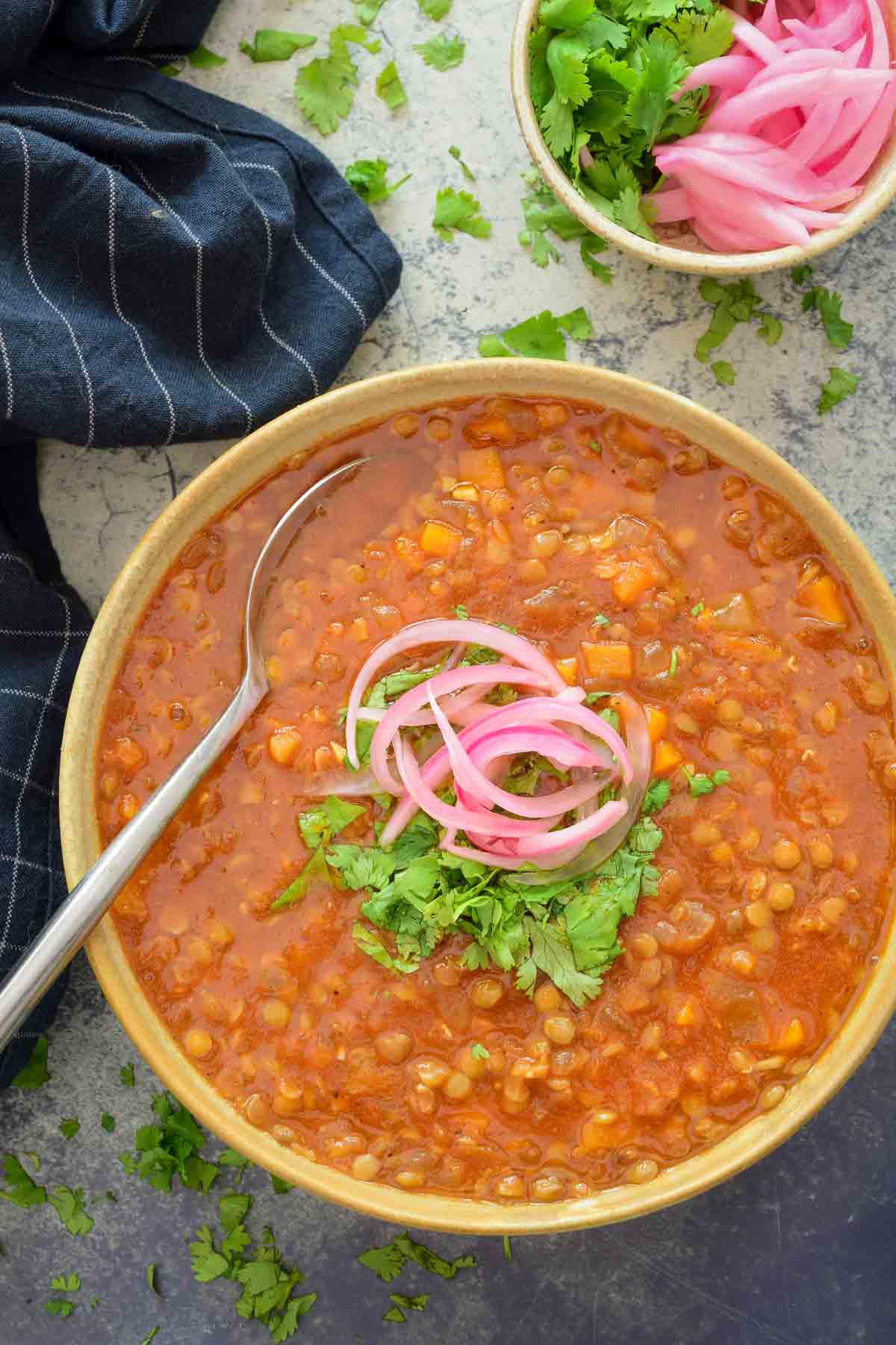 An overhead photo of a beige ceramic bowl is filled with Moroccan lentil soup and is garnished with cilantro and pickled red onions. A spoon sits in the back of the bowl.