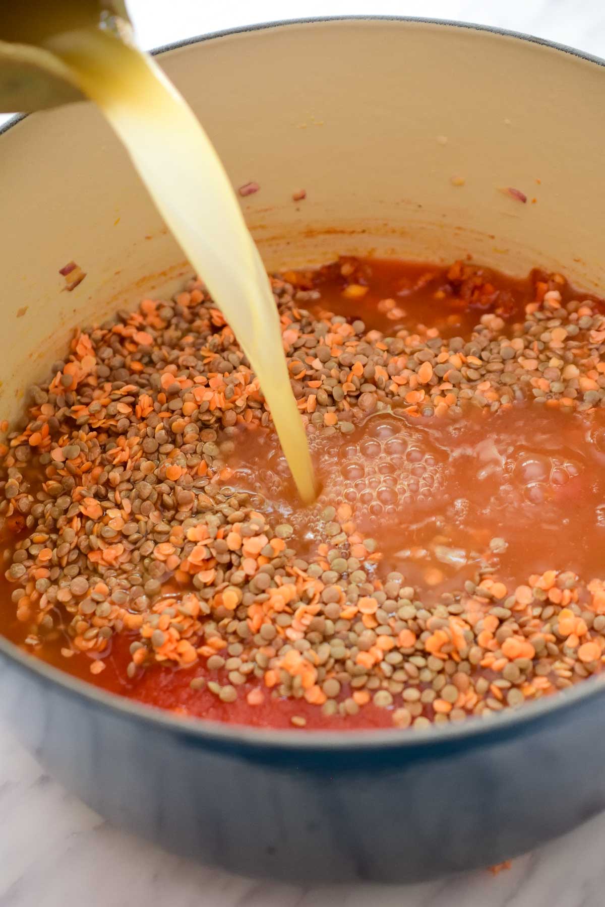 Inside a blue dutch oven that has lentils and tomato puree. Vegetable stock is being poured over the lentils into the pot.
