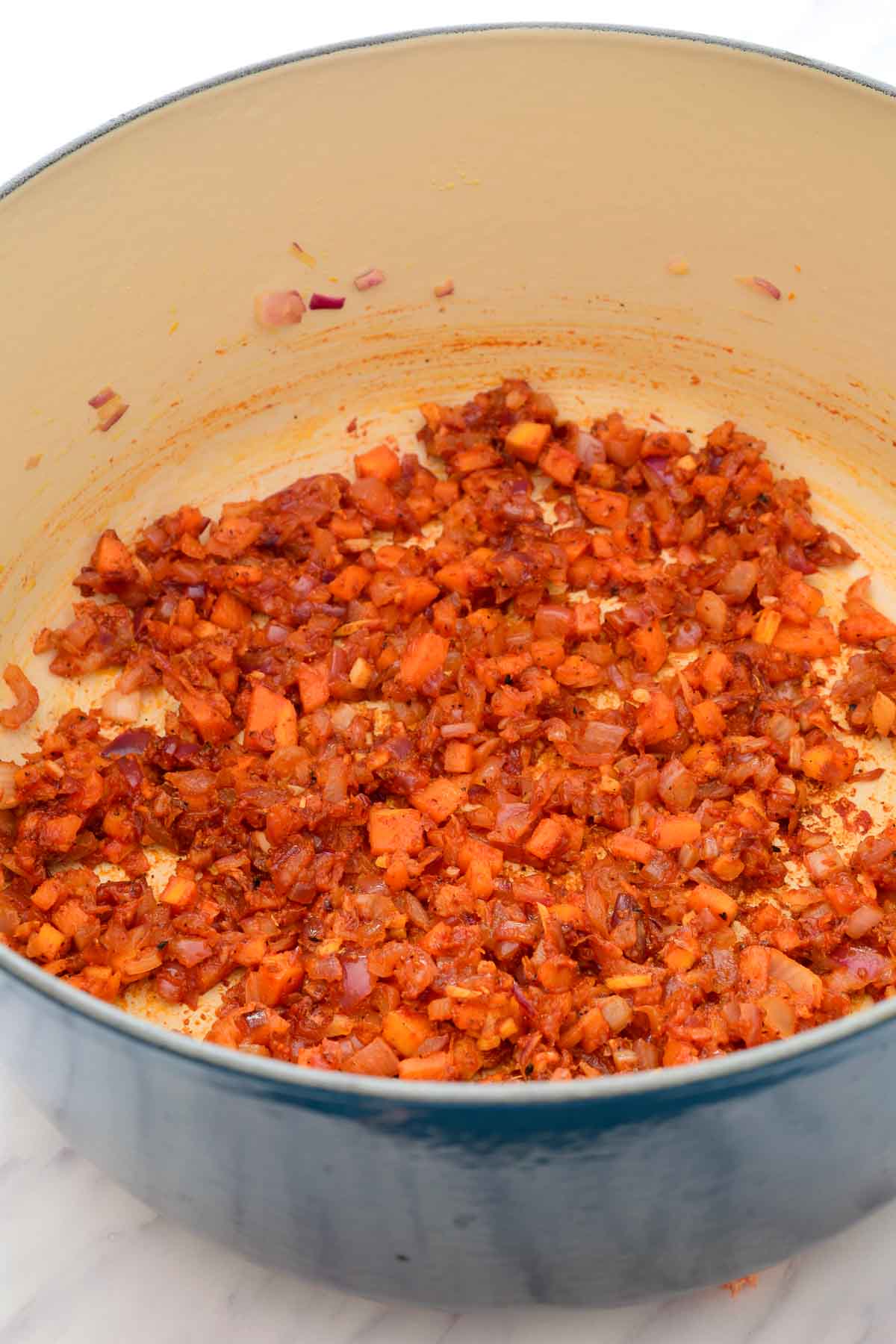 A blue dutch oven with fried onion, carrot, ginger and garlic that is covered in tomato paste.