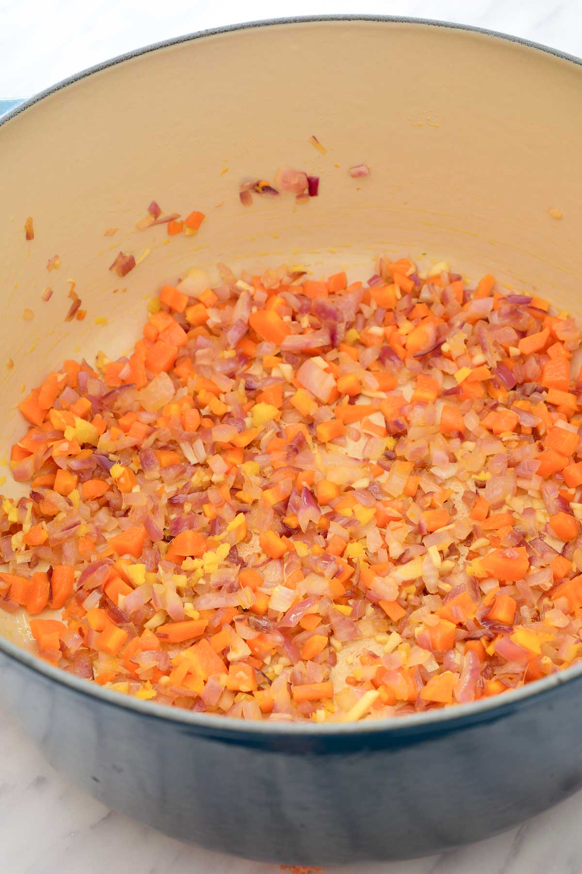 A blue dutch oven with fried onion, carrot, ginger and garlic on the bottom.