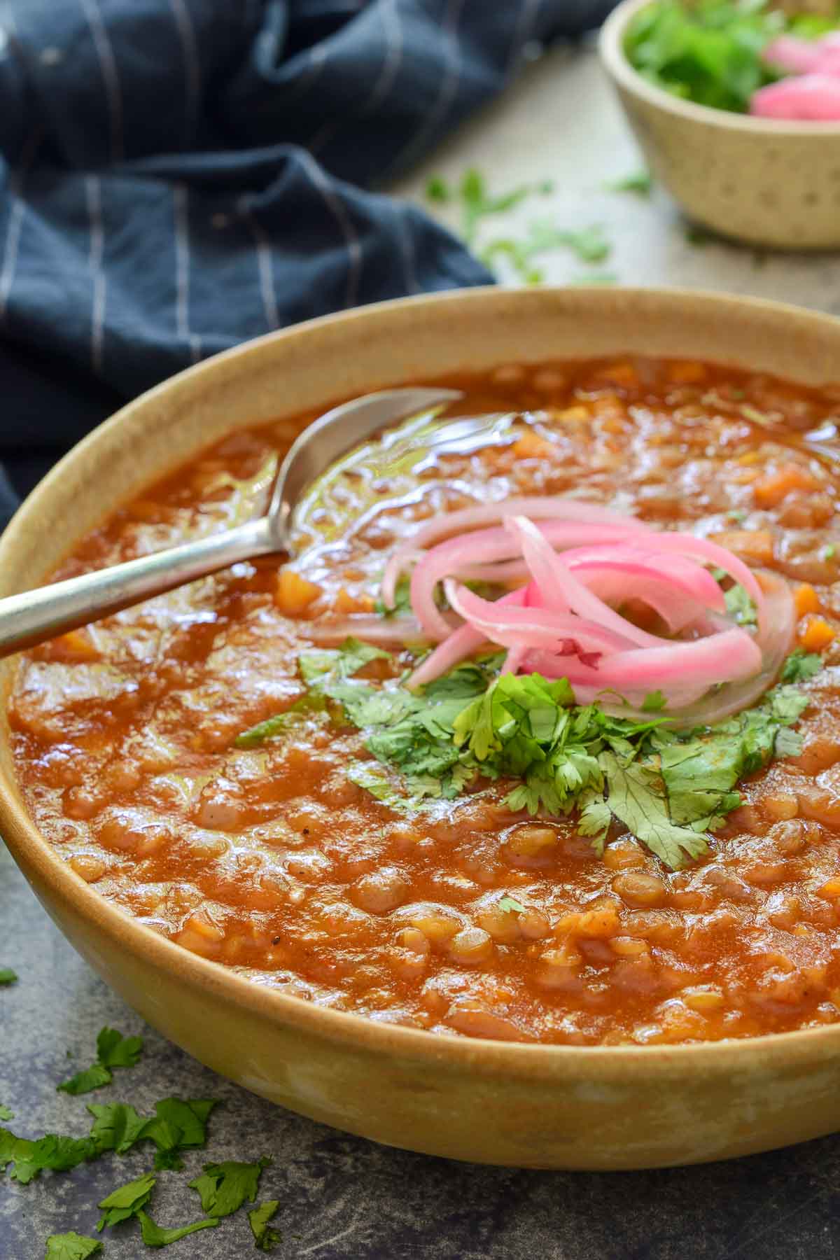 A 45-degree angle of a beige ceramic bowl is filled with Moroccan lentil soup and is garnished with cilantro and pickled red onions. A spoon sits in the back of the bowl.