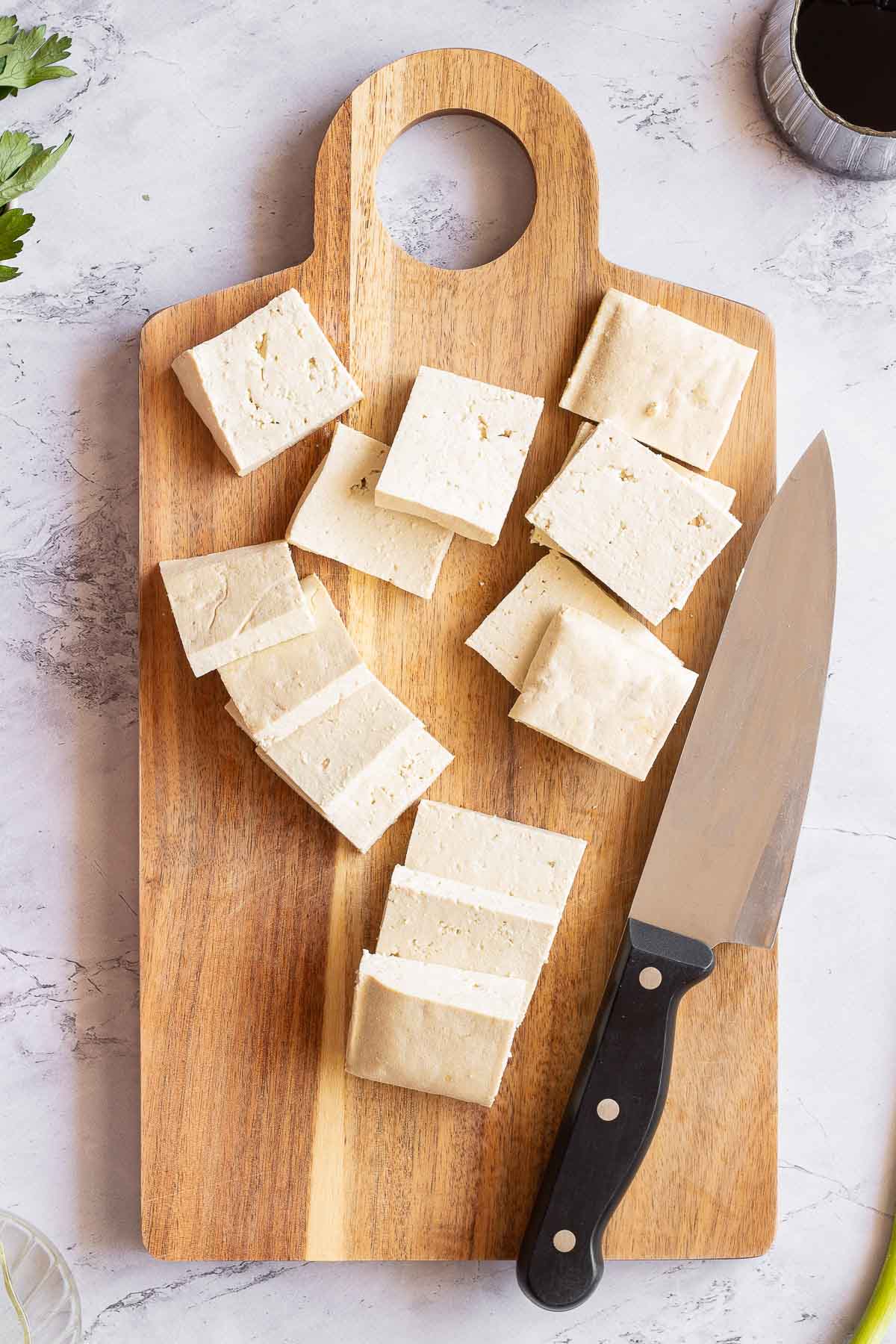 Slices of firm tofu and a kitchen knife sit on a wooden cutting board atop a white marble surface.