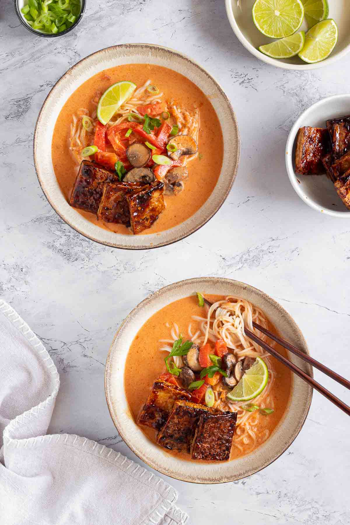 Two ceramic bowls filled with Thai red curry ramen on a white marble surface. The bottom bowl also has wooden chopsticks in it. Smaller white bowls of seasoned tofu, lime slices and chopped green onion surround the bowls. A white napkin lies in the bottom left corner.