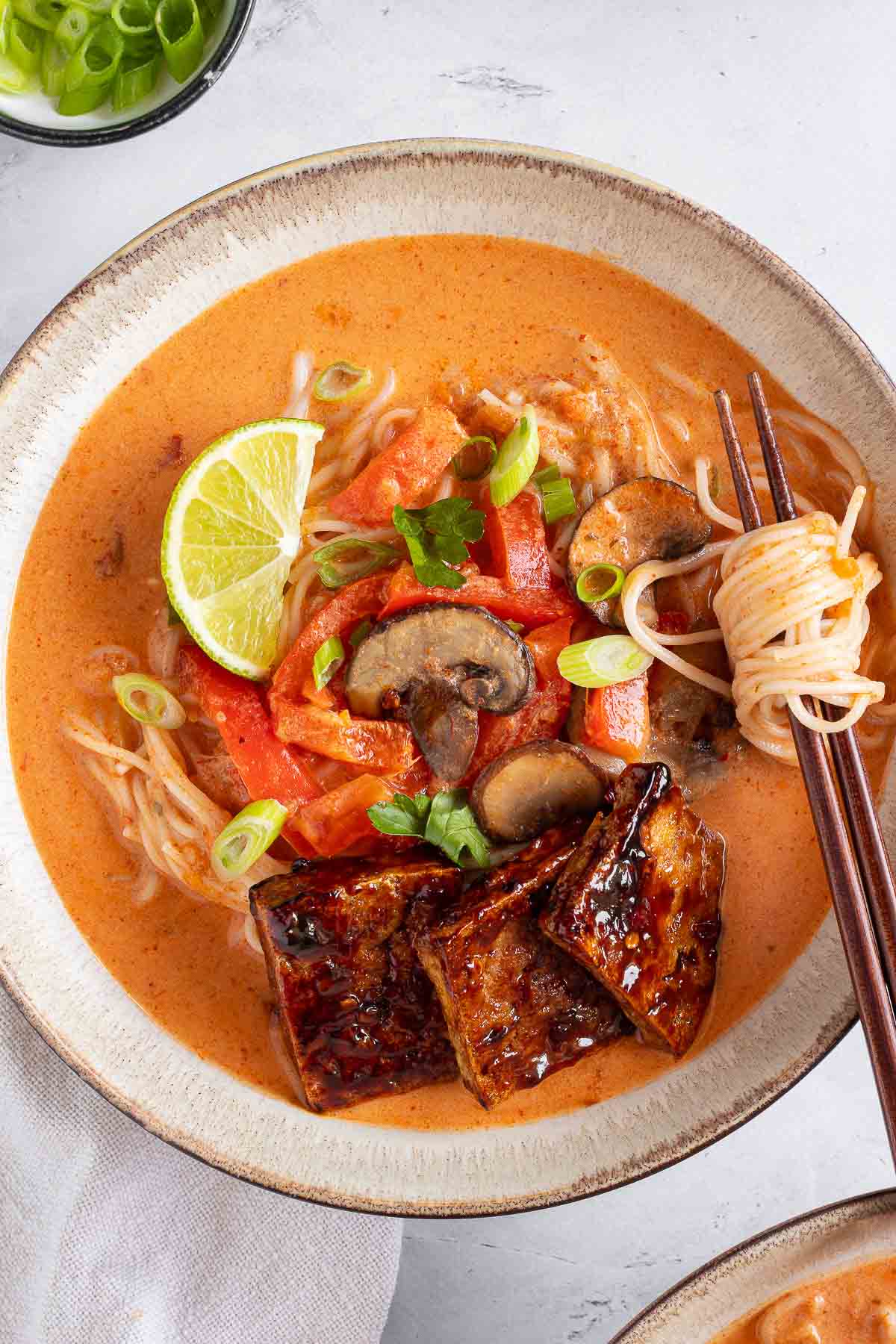 A close-up photo of Thai red curry ramen noodles in a light-colored ceramic bowl. Some ramen noodles are wrapped around a pair of wooden chopsticks sitting on the side of the bowl.