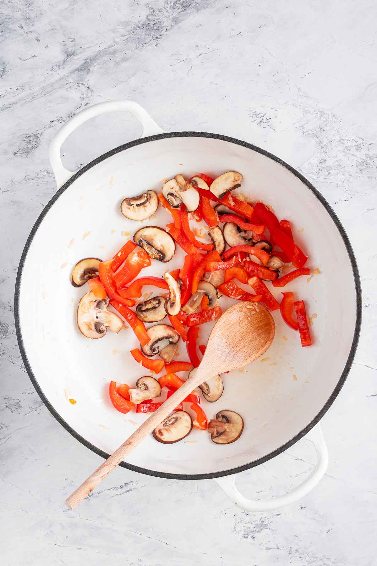 A large white pot sitting on a white marble surface contains fried slices of red pepper and mushrooms. A wooden spoon is resting in the pot.