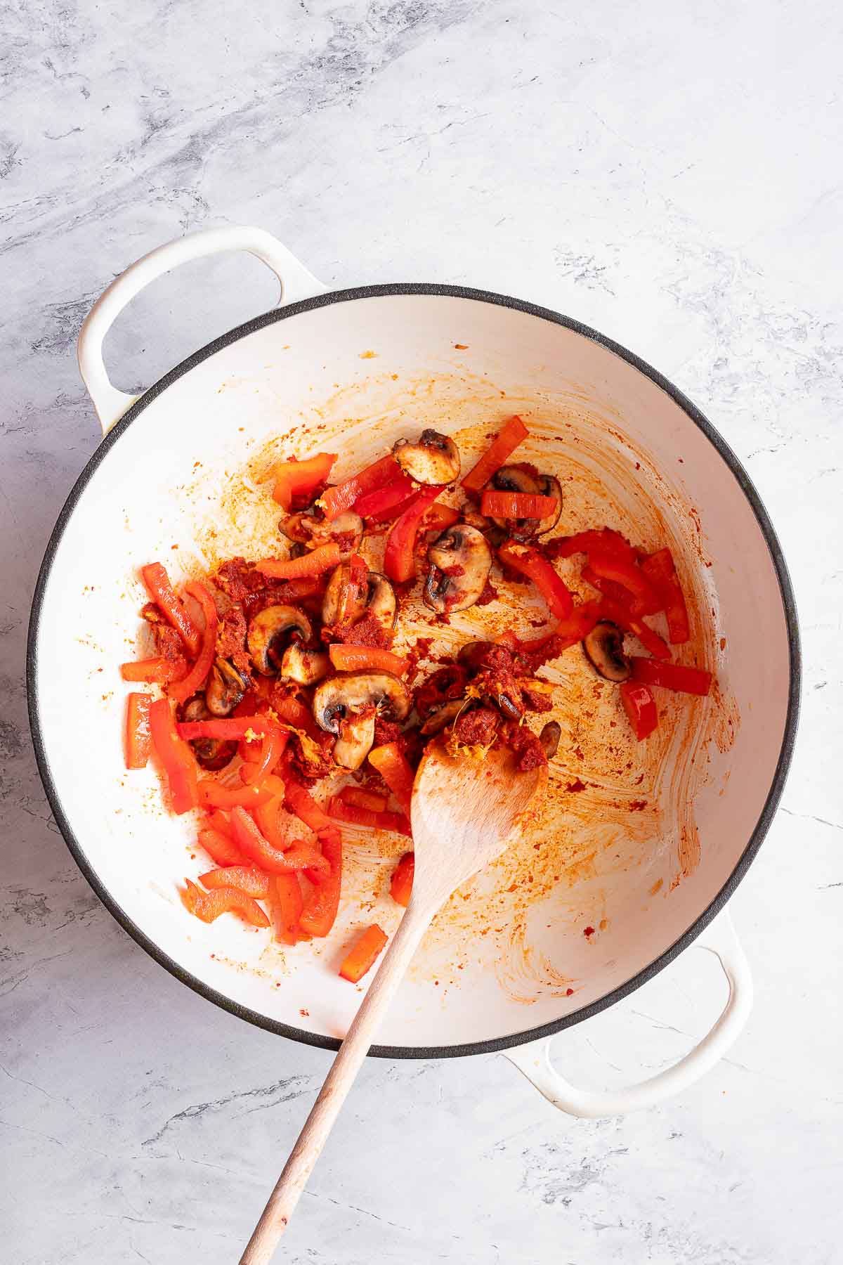 A large white pot sitting on a white marble surface contains fried slices of red pepper and mushrooms seasoned with red curry paste. A wooden spoon is resting in the pot.