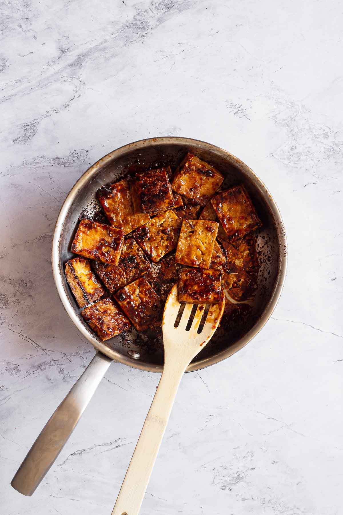 A stainless steel pan is filled with dark brown tofu glazed with a sticky sauce. There is a wooden spoon under one of the tofu slices.