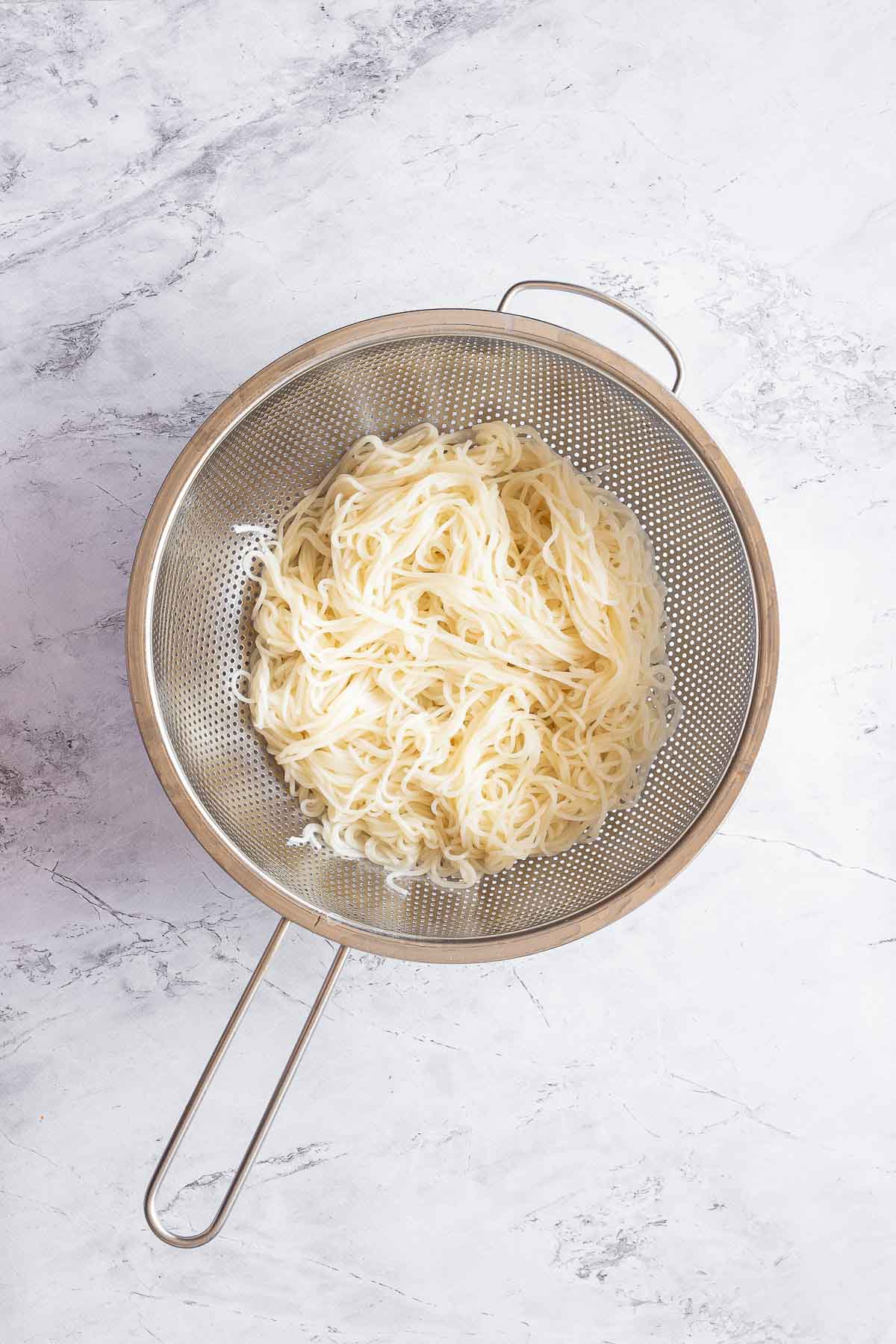 A strainer filled with cooked ramen noodles sits on a white marble surface.
