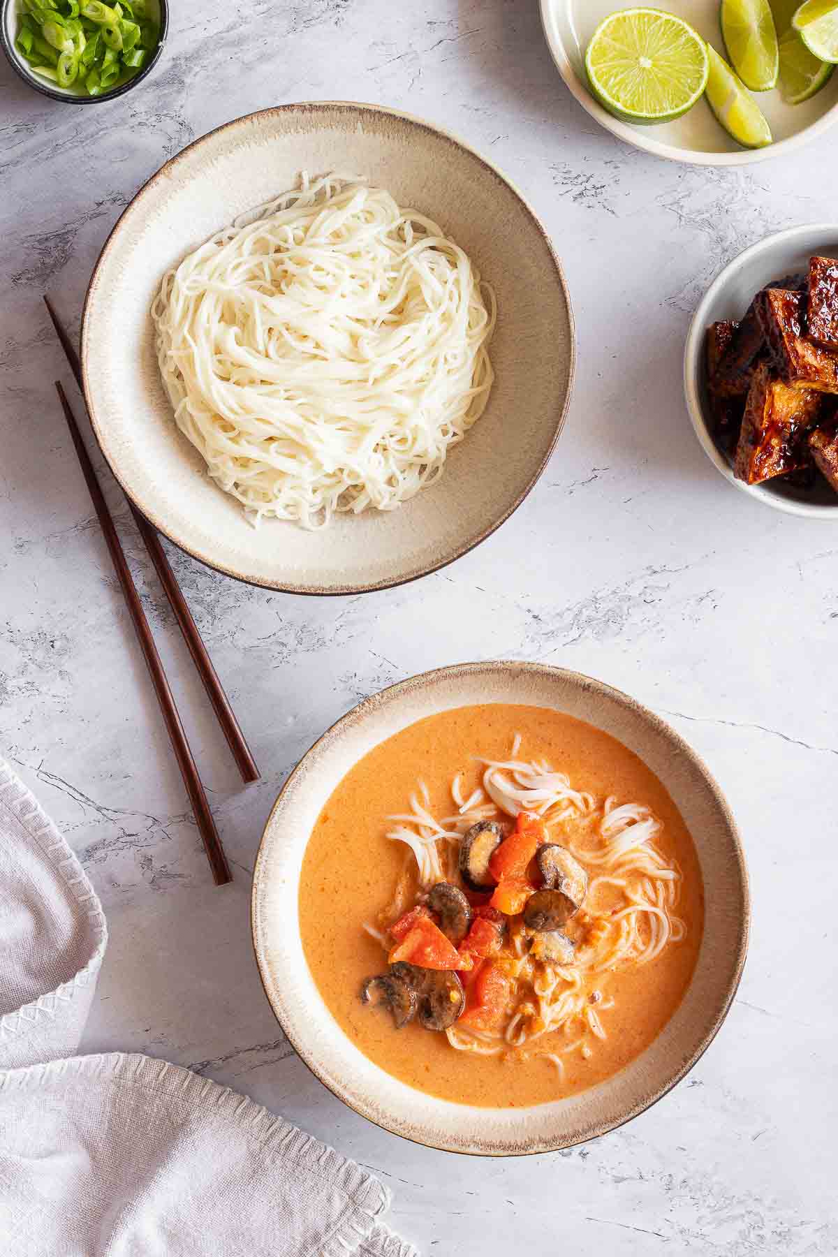 A ceramic bowl contains ramen noodles and Thai curry broth with vegetables. A second bowl contains plain ramen noodles. A third bowl contains seasoned tofu. Wooden chopsticks and a white napkin surround the bowls sitting on a white marble surface.